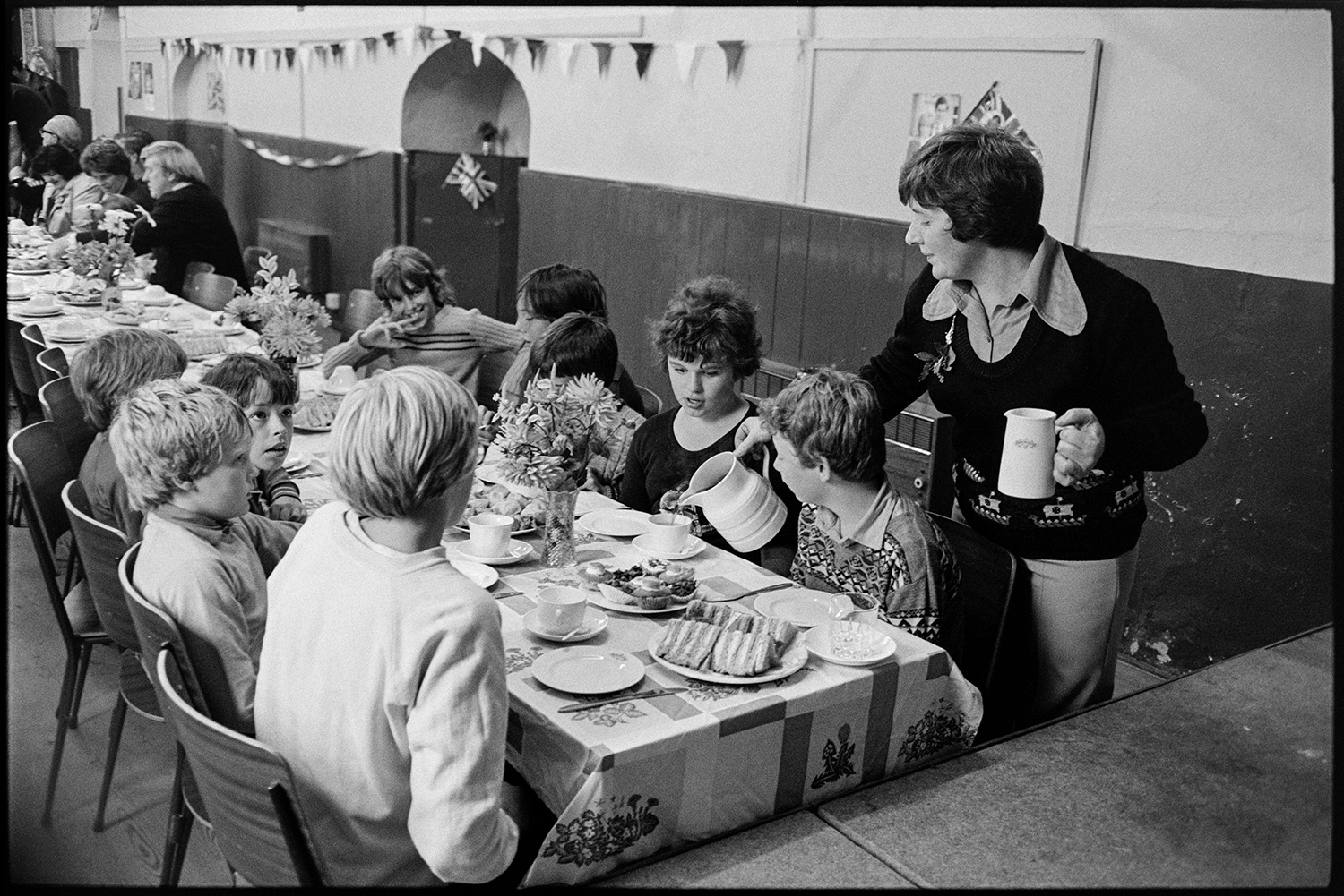 Village sports day, speeches, eating tea in hall, races, people chatting. 
[Children having a tea of sandwiches and cakes in Roborough Village Hall after Roborough Sports Day. A woman is pouring them drinks. The hall is decorated with bunting and flags.]