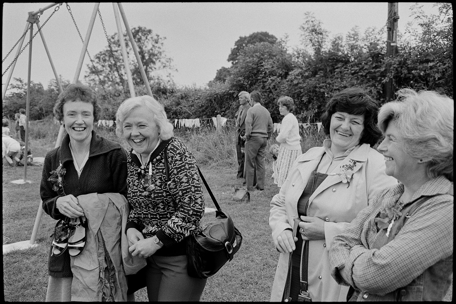 Village sports day, women, chatting, making raffle draw. 
[Four women talking and laughing in a park at Roborough Sports Day. Swings can be seen in the background.]