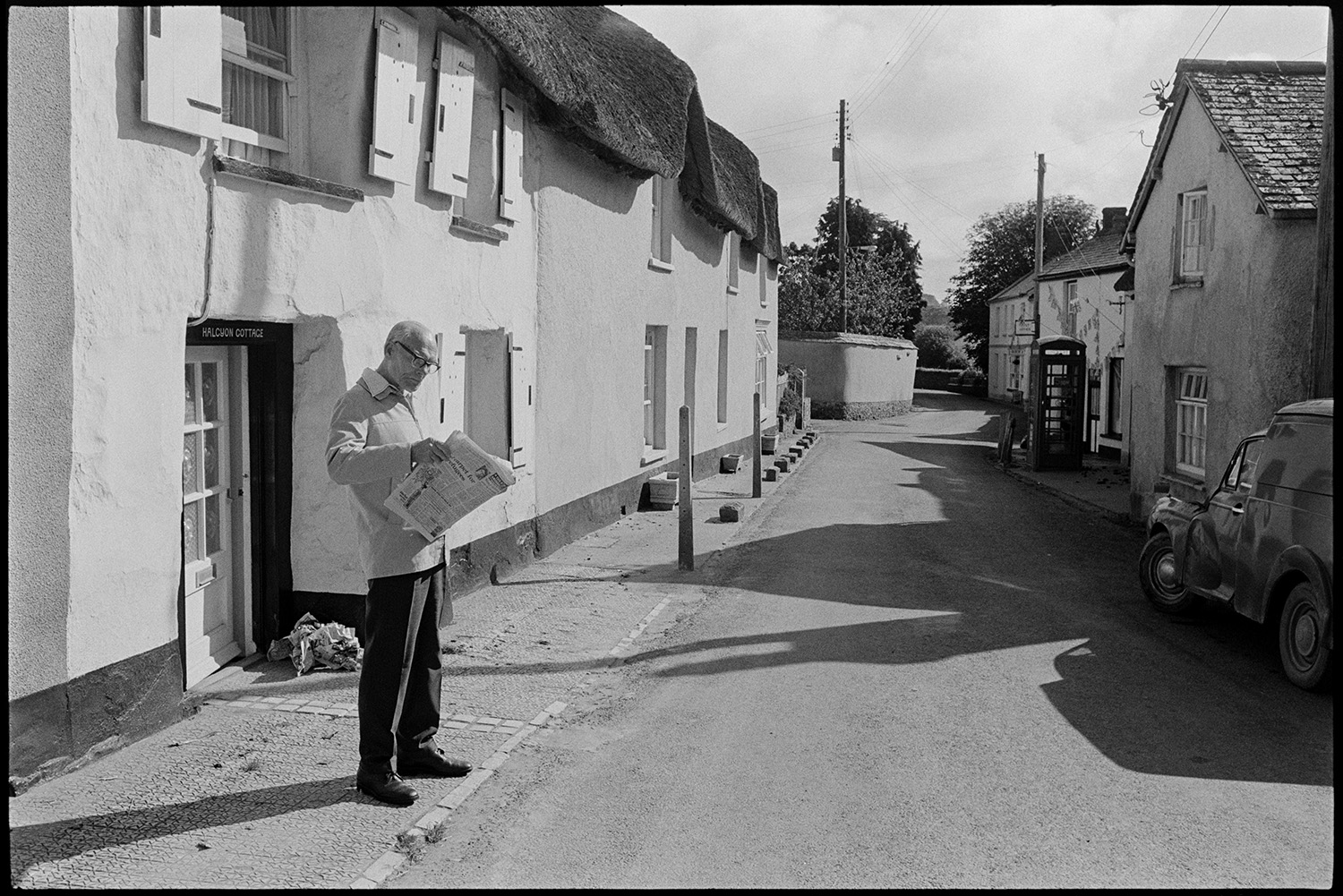Street scene with man reading newspaper. 
[A man reading a newspaper outside a thatched cottage with window shutters in Fore Street, Dolton. A van is parked in the road opposite him and a telephone box can be seen further down the street.]