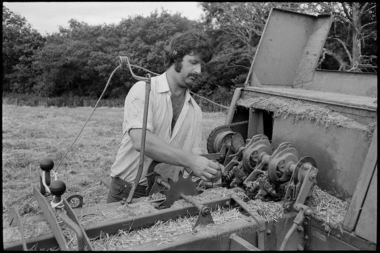 Engineer mending baler. 
[A man fixing the mechanism of a baler in a field at Densham, Ashreigney.]