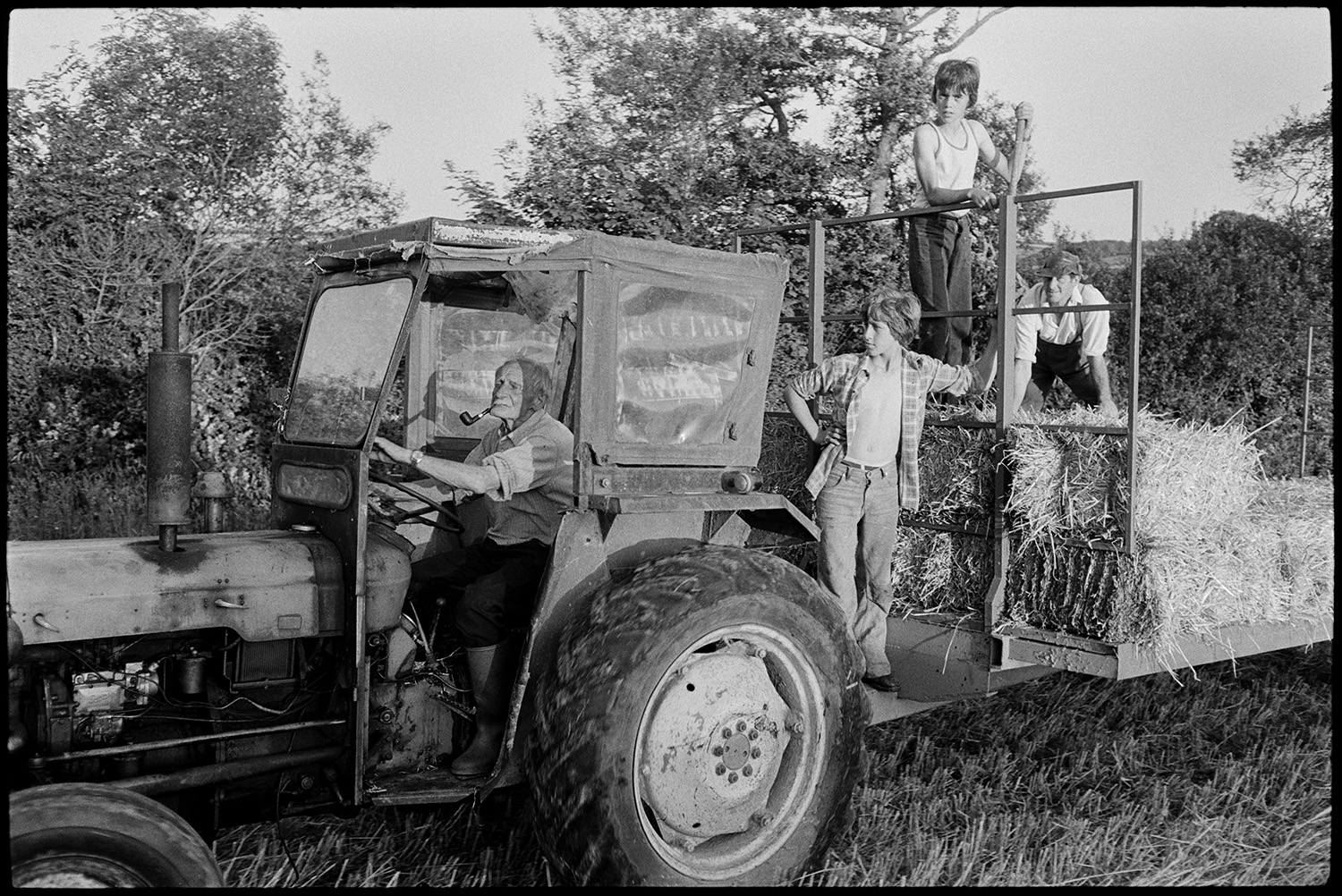 Combine harvester, farmers loading straw bales onto trailer. 
[A man driving a tractor and trailer loaded with straw bales in a field at Hollocombe. He is smoking a pipe. Another man and two boys are stood on the back of the trailer waiting to collect the next bales.]