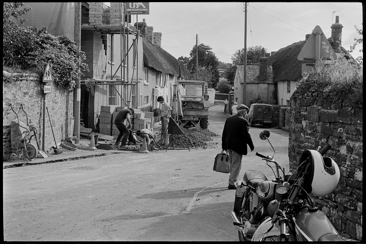 Street scenes, woman sweeping pavement. 
[Men digging up the road outside a house with scaffolding in Fore Street, Dolton. A man carrying a bag is walking by and a motorbike is parked in the foreground.]