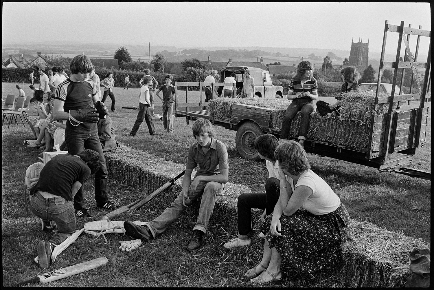 Spectators at cricket match sitting on straw bales, church tower in background, tractors. 
[Teenagers sat on straw bales at a cricket match in a field at Iddesleigh. One man is either putting on or removing knee pads for batting. Two girls are sat on straw bales on a trailer in the background and Iddesleigh church tower can be seen in the distance.]