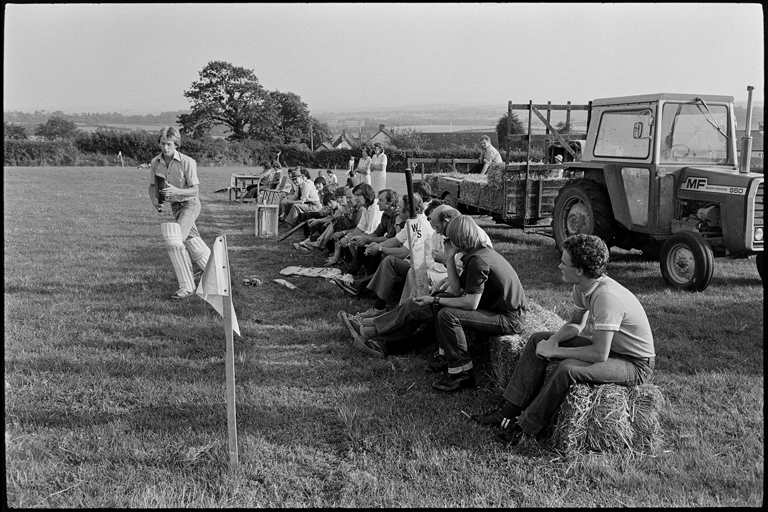 Spectators at cricket match sitting on straw bales, church tower in background, tractors. 
[Spectators sat on straw bales watching a cricket match in a field at Iddesleigh. A man is walking up to go into bat. He is wearing knee pads and holding a cricket bat. A tractor and trailer with more straw bales is parked behind the spectators.]