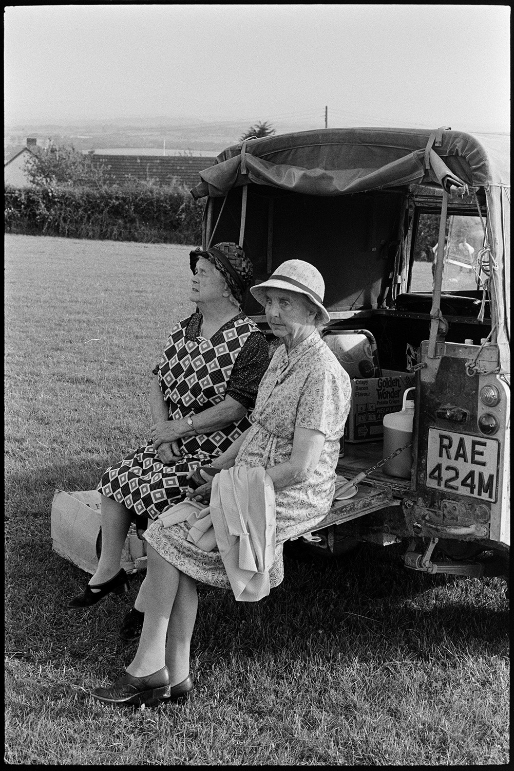 Spectators at cricket match sitting on straw bales, church tower in background, tractors. 
[Two women sat on the back of a Land Rover, watching a cricket match in a field at Iddesleigh.]