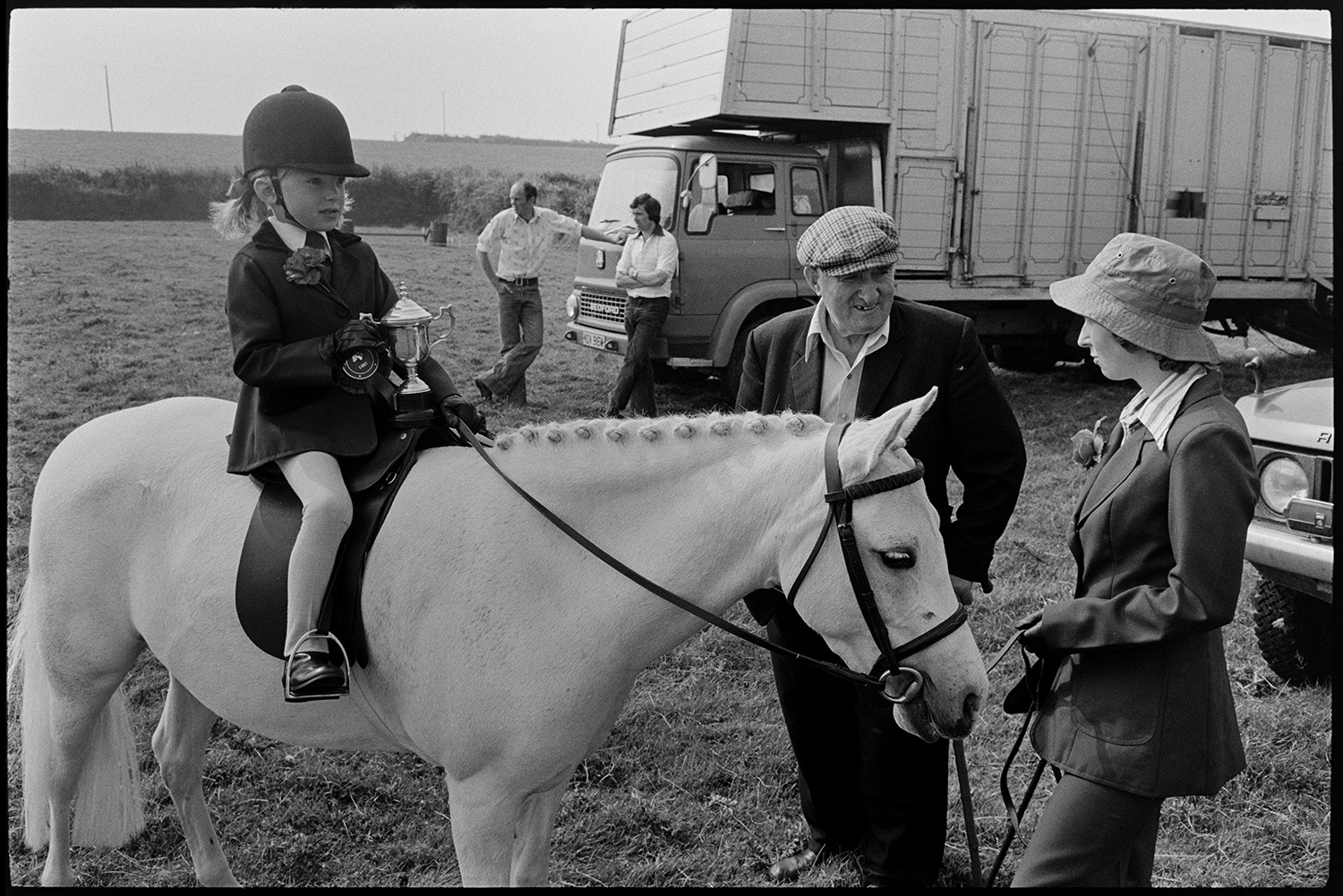 Competitors preparing gymkhana, horseboxes. 
[A young girl on a horse with a plaited mane at Beaford Gymkhana. She is holding a cup she has won. A woman is holding the reins of the horse while talking to a man. Two people are leaning against a horsebox or animal transporter in the background.]