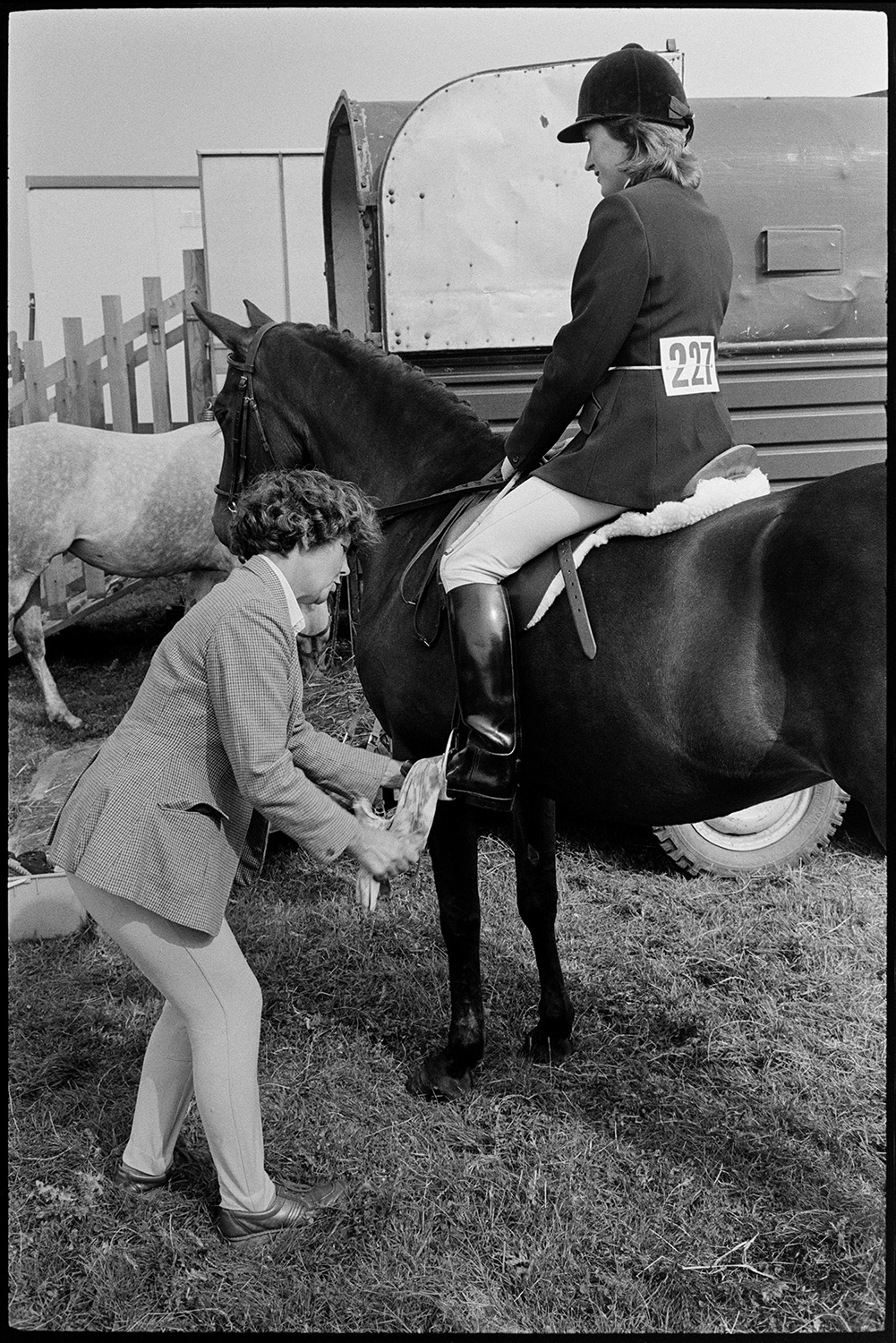 Competitors preparing gymkhana, horseboxes. 
[A woman shining the boots of a mounted horse rider at Beaford Gymkhana, by a horsebox.]