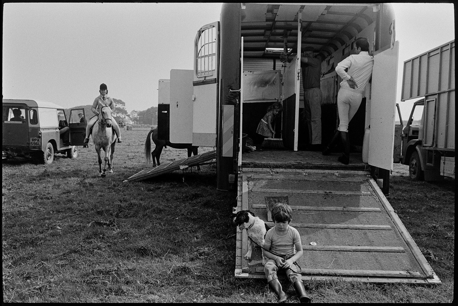 Competitors preparing gymkhana, horseboxes. 
[A young child and a dog sitting on the ramp leading up into a horsebox at Beaford Gymkhana. A  competitor is riding towards them on horseback.]