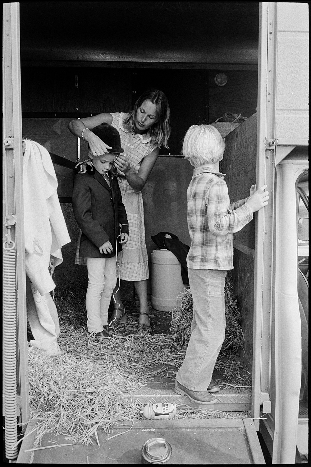 Competitors preparing gymkhana, horseboxes. 
[A woman in a horsebox putting a riding hat on a child, ready to compete in the Beaford Gymkhana. Another child is watching them.]