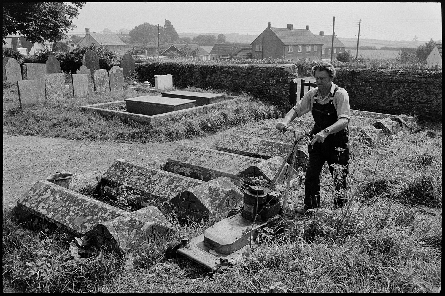 Man mowing grass in graveyard. Gravestones. 
[A man cutting the grass between gravestones in Beaford churchyard, using a lawn mower.]