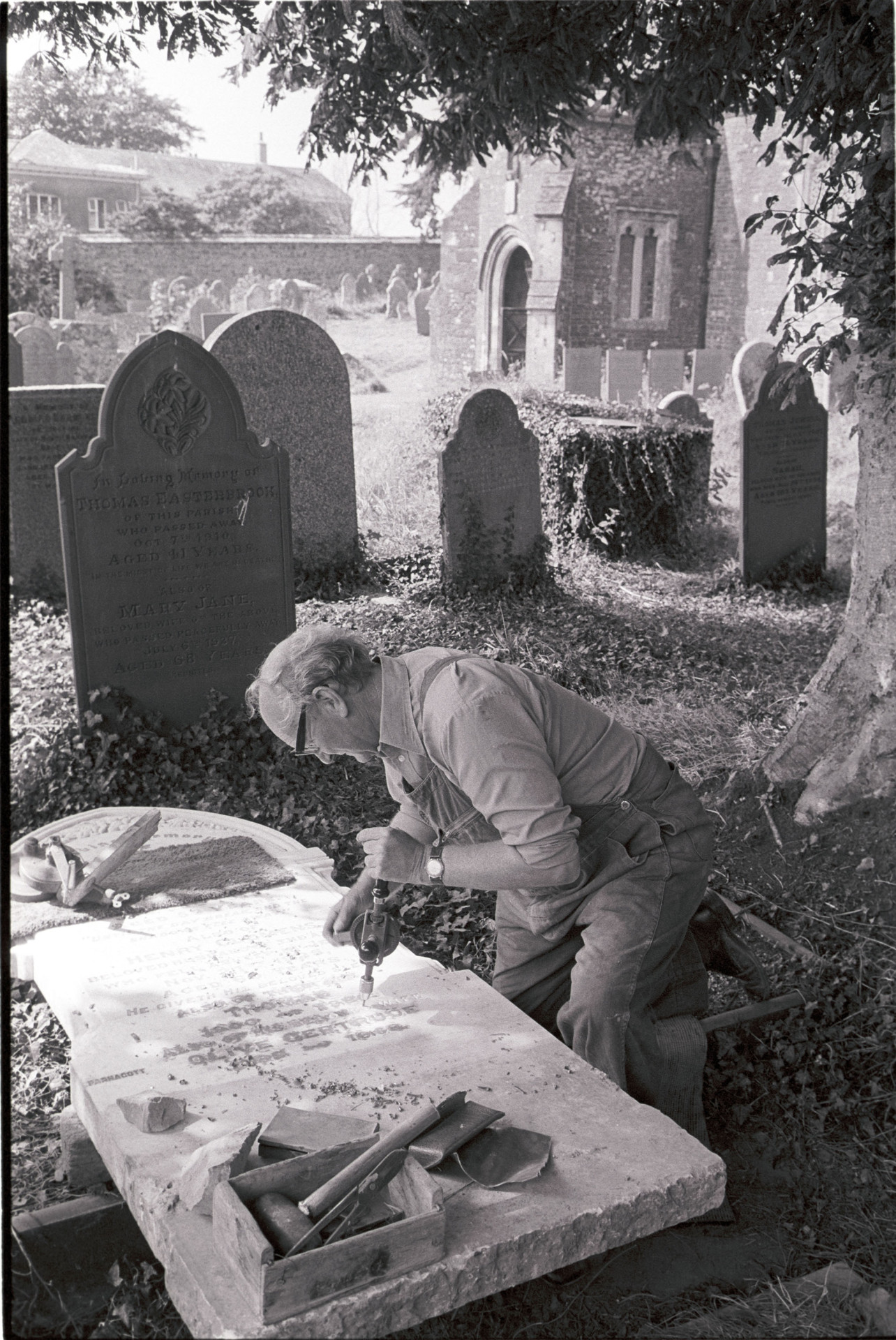 Man working on gravestone, this had been worked by his father and his boss before him. 
[Bruce Edyvean, Monumental Mason, adding an inscription to a gravestone in Beaford churchyard, using a hand drill. Other gravestones and the church porch are visible in the background.]