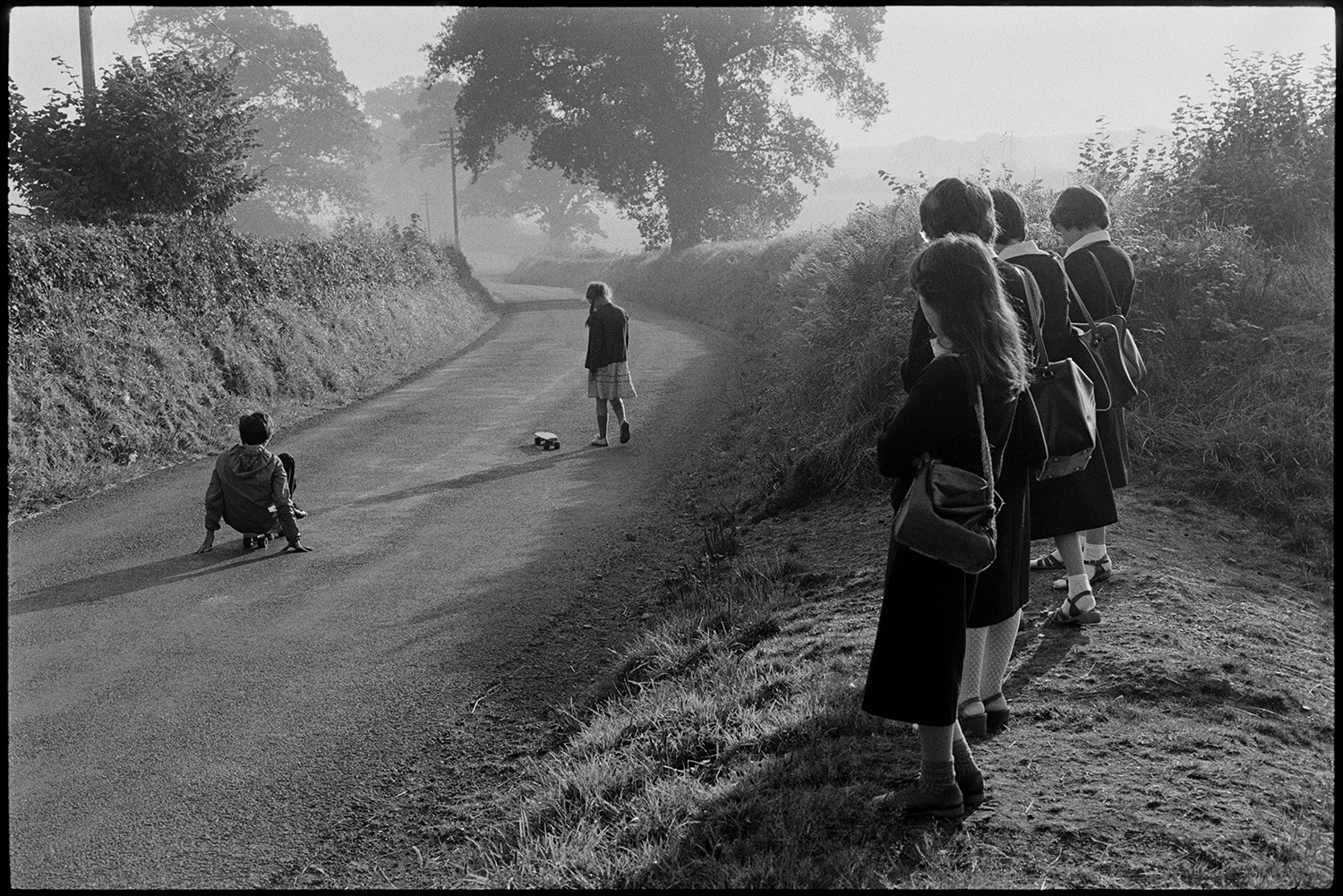 Children waiting for school bus, under oak tree, morning. Wearing uniform. 
[Six children in school uniform waiting for the school bus on the verge of a road at Lovistone, Merton. Two of the children are playing on skateboards in the road.]
