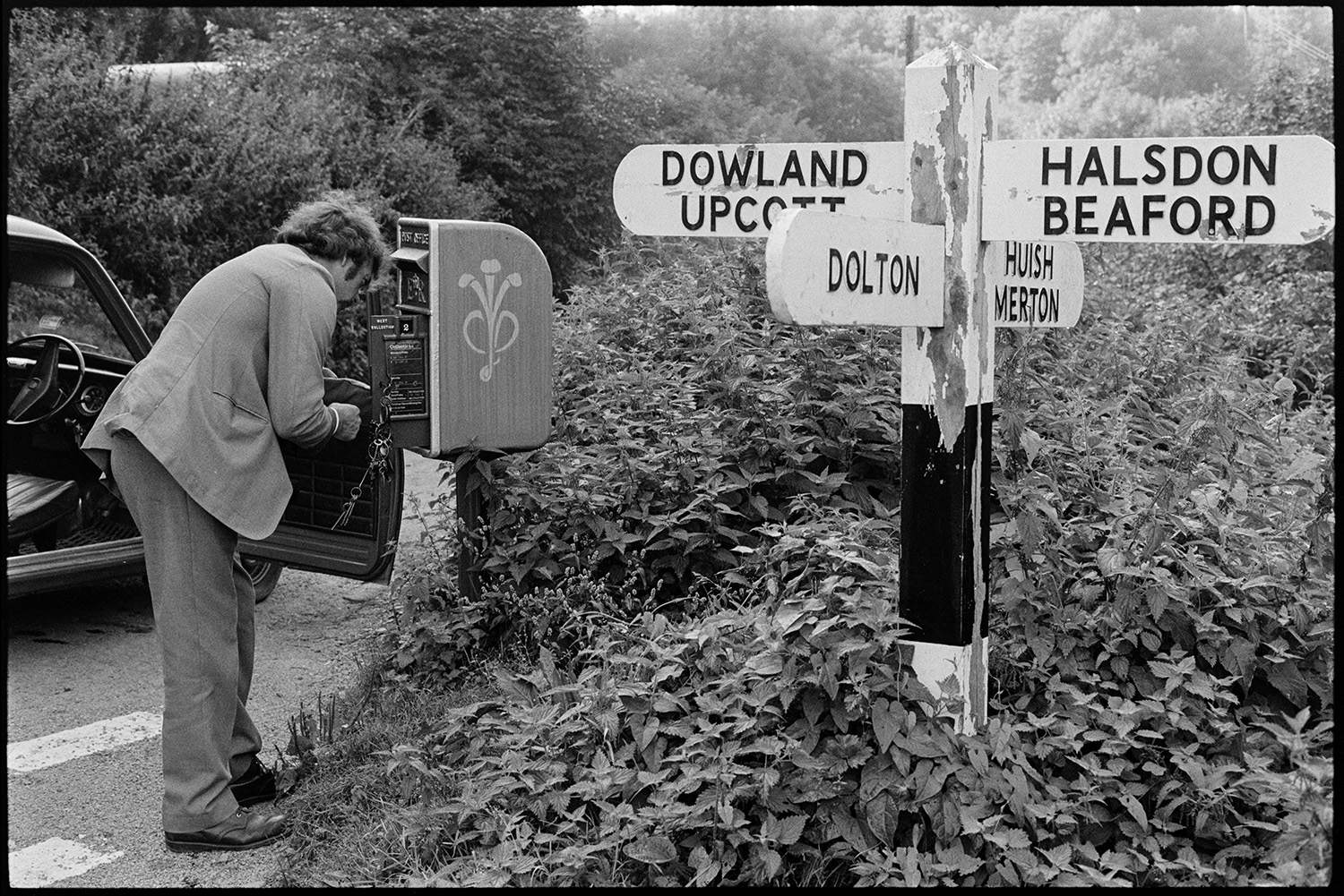 Portman emptying letter box next to road sign, nettles. 
[A postman emptying a post box at Woolridge Cross, Dolton, next to a signpost overgrown by stinging nettles.]