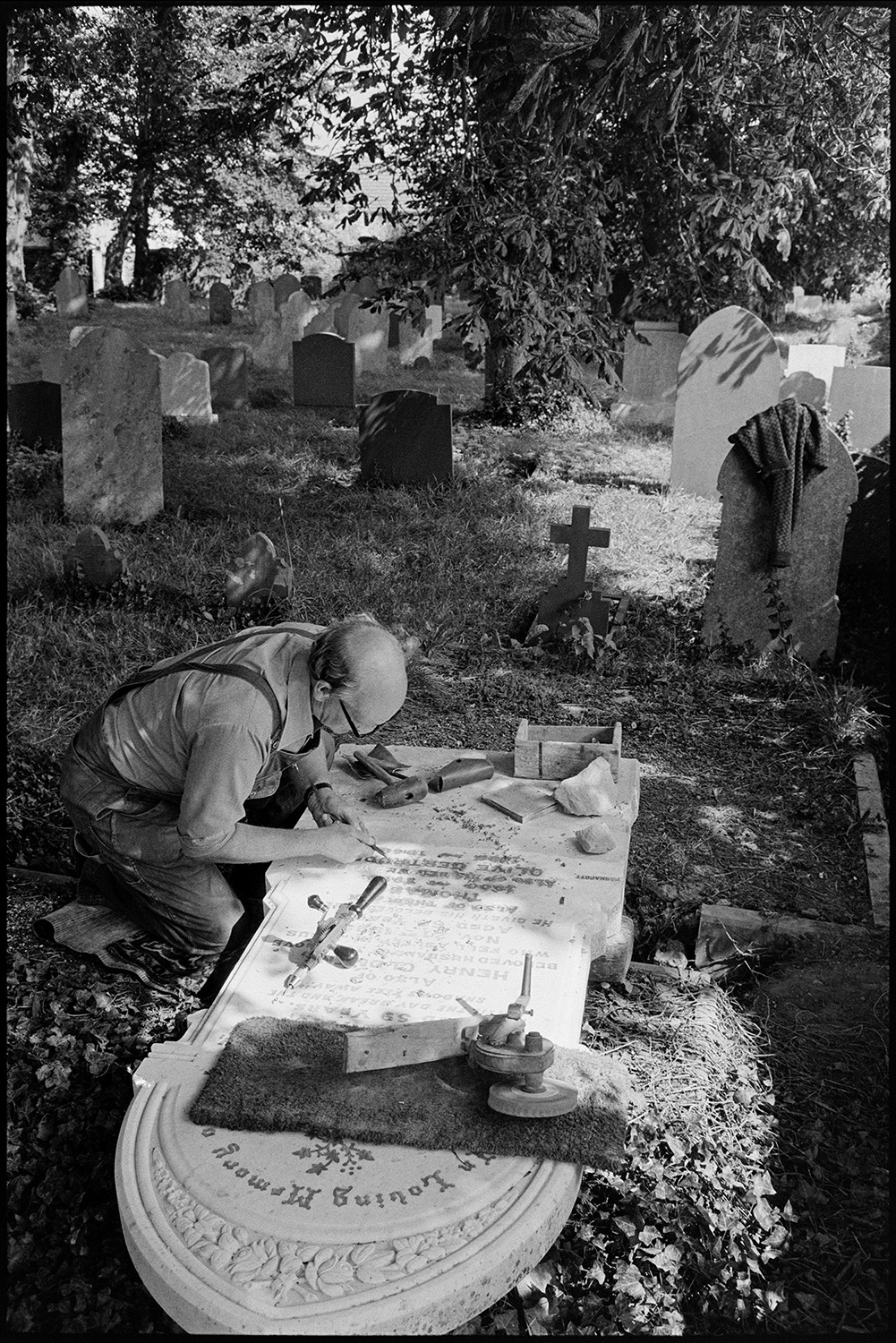 Man working on gravestone, this had been worked by his father and his boss before him. 
[Bruce Edyvean engraving a gravestone in Beaford Churchyard. He is using various tools including a hand drill and a chisel. His father and his father's boss had both worked on the same stone.]