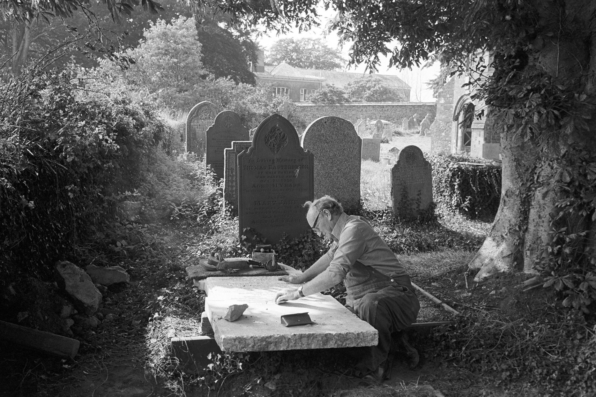 Man working on gravestone, this had been worked by his father and his boss before him. 
[Bruce Edyvean, Monumental Mason, adding an inscription to a gravestone in Beaford churchyard, under a tree. His tools, including a hand drill, are resting on the gravestone. Other gravestones and the church porch are visible in the background.]