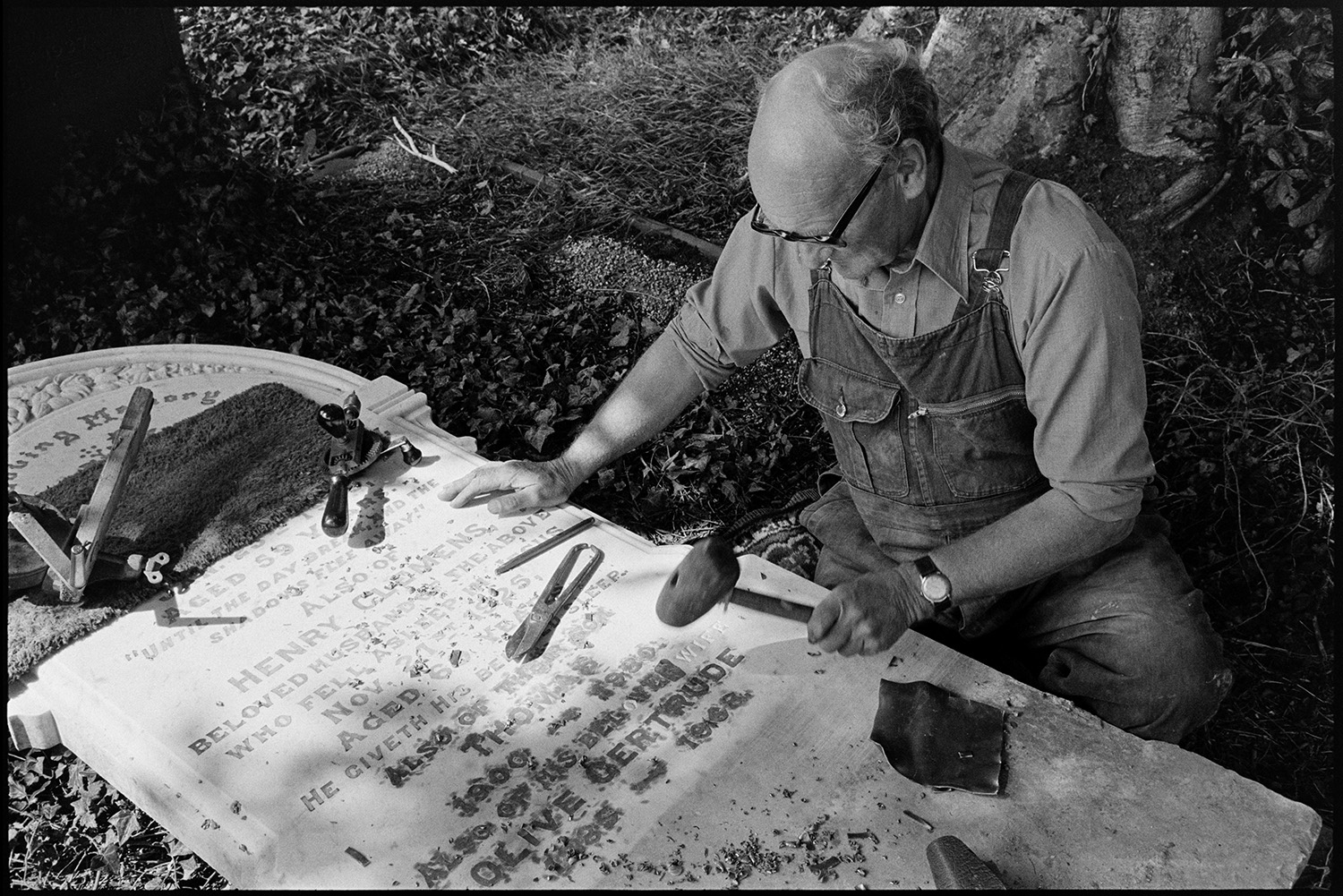 Man working on gravestone, this had been worked by his father and his boss before him. 
[Bruce Edyvean engraving a gravestone in Beaford Churchyard. He is using various tools including a hand drill and a mallet. His father and his father's boss had both worked on the same stone.]