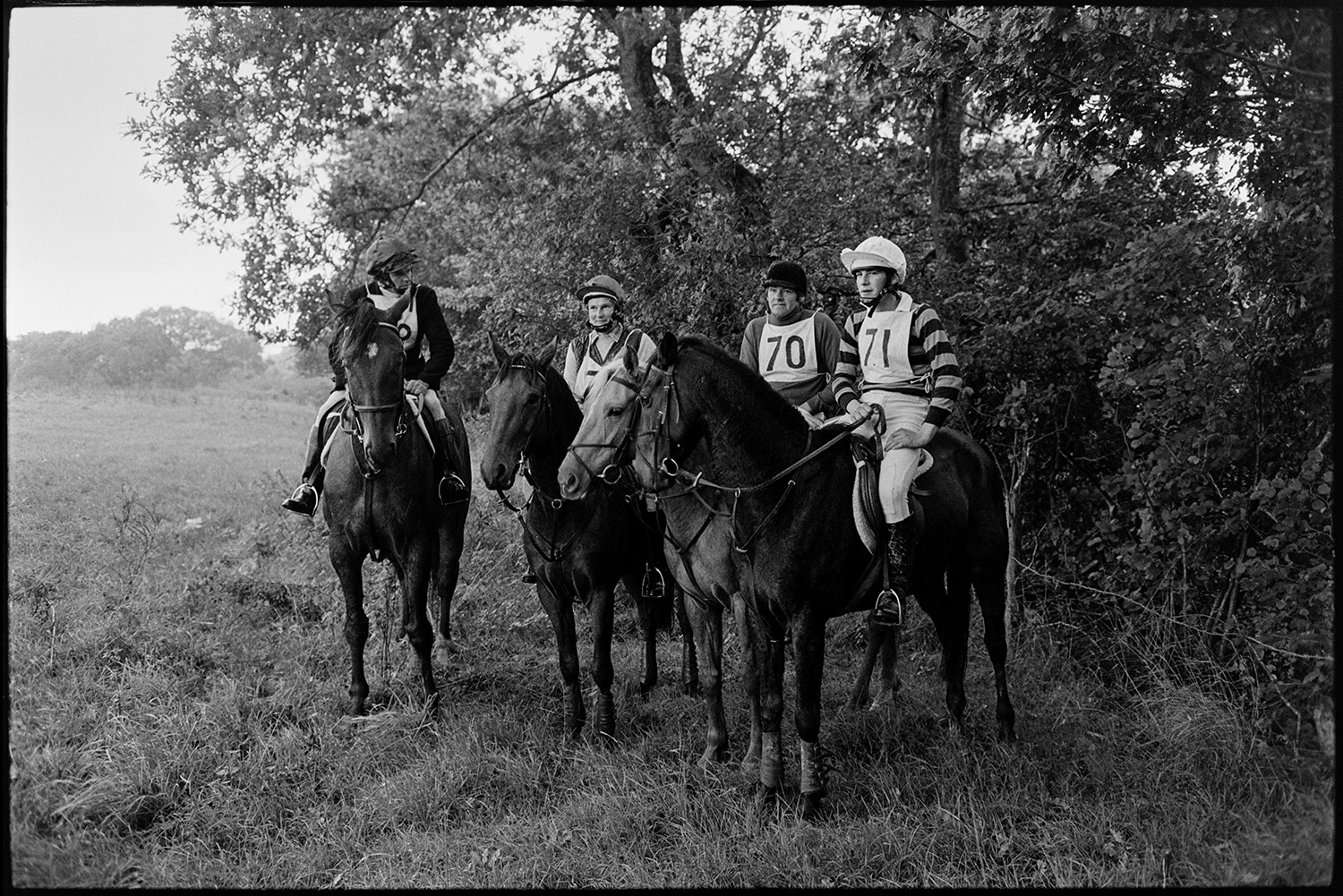 Competitors with horses at Steeplechase. 
[Four mounted horse riders sheltering under trees by a hedge at a steeplechase at Winkleigh Aerodrome.]