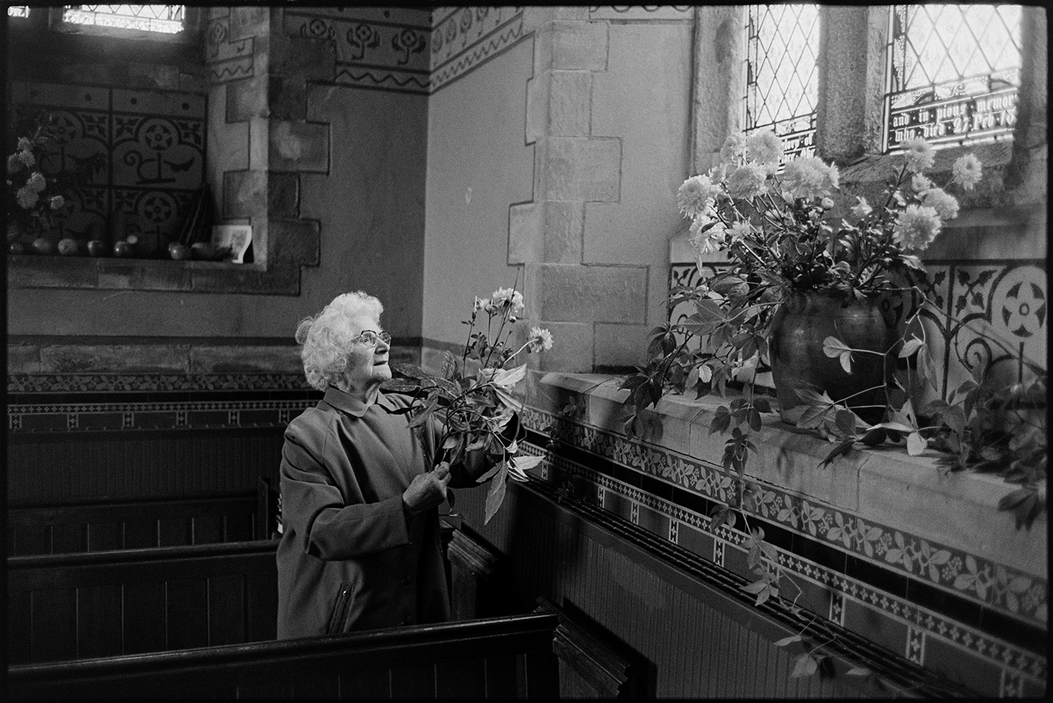 Women putting flowers in church for harvest festival. <br />
[A woman holding flowers and reaching up to put them in an arrangement in a large vase on a tiled windowsill for the Harvest Festival at Winkleigh Church.]