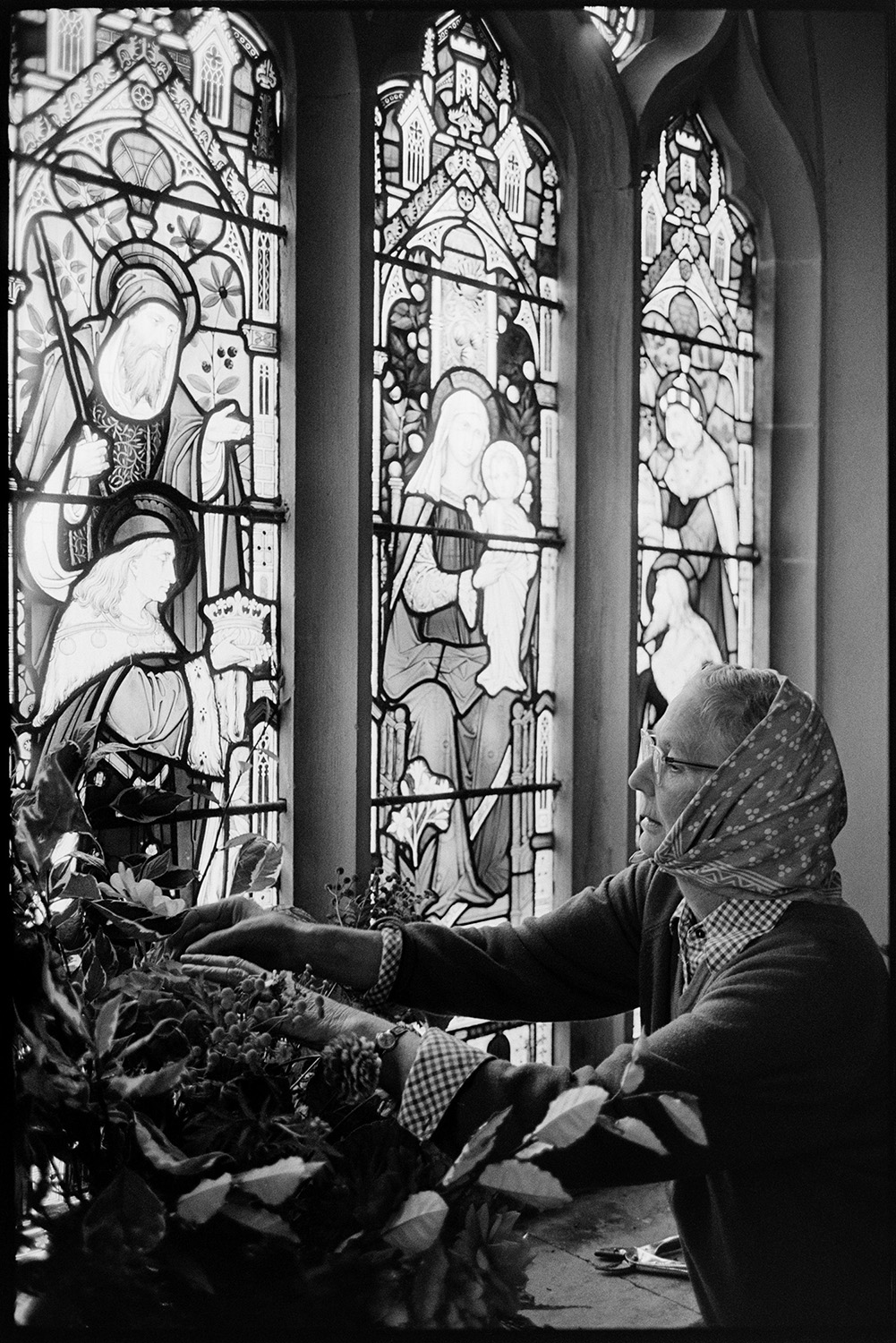 People decorating church for Harvest Festival.
[A woman placing flowers in an arrangement on the windowsill of a stained glass window for the Harvest Festival at Dolton Church. She is wearing a headscarf]