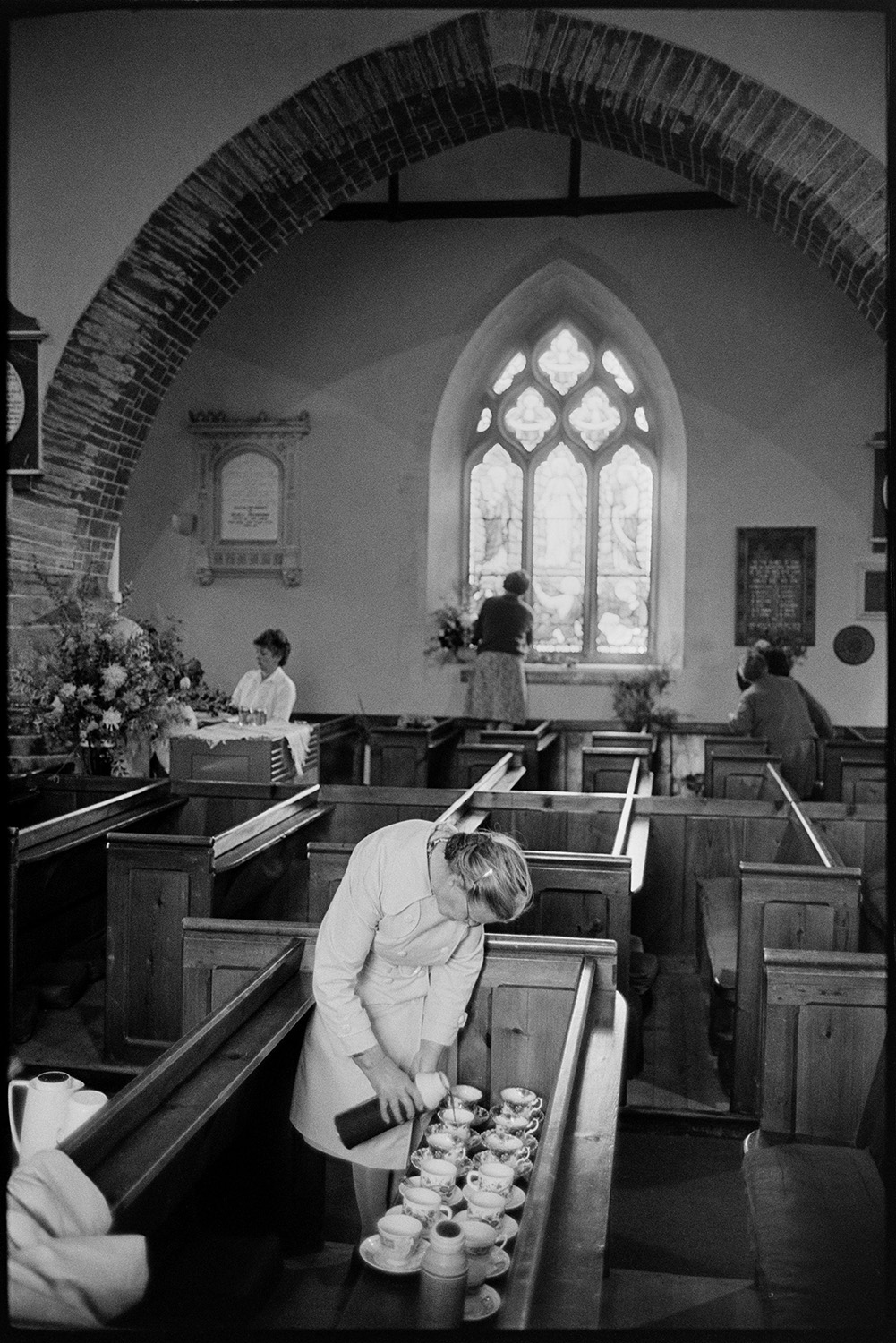 People decorating church for Harvest Festival.
[A woman pouring drinks from a flask into cups laid out on a pew, for the others who are decorating Dolton Church for the Harvest Festival with flower arrangements.]