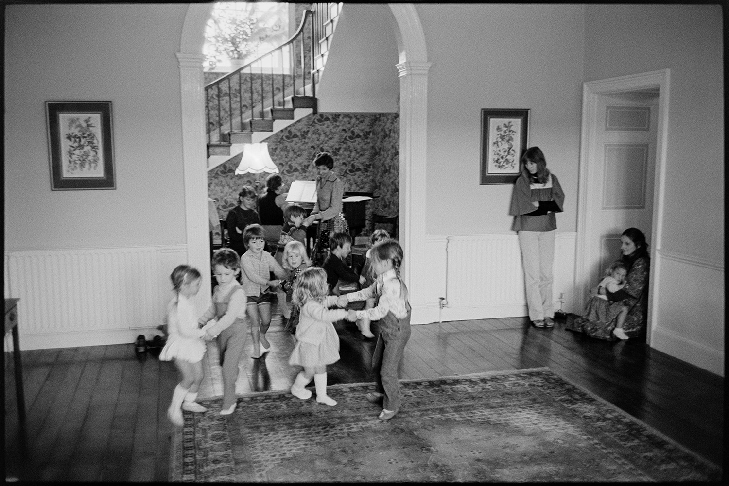 Children's dance class in large country house.
[Young children dancing at Marwood House by an archway, with women watching. Another woman is playing a piano in the background. A staircase can be seen through the archway.]