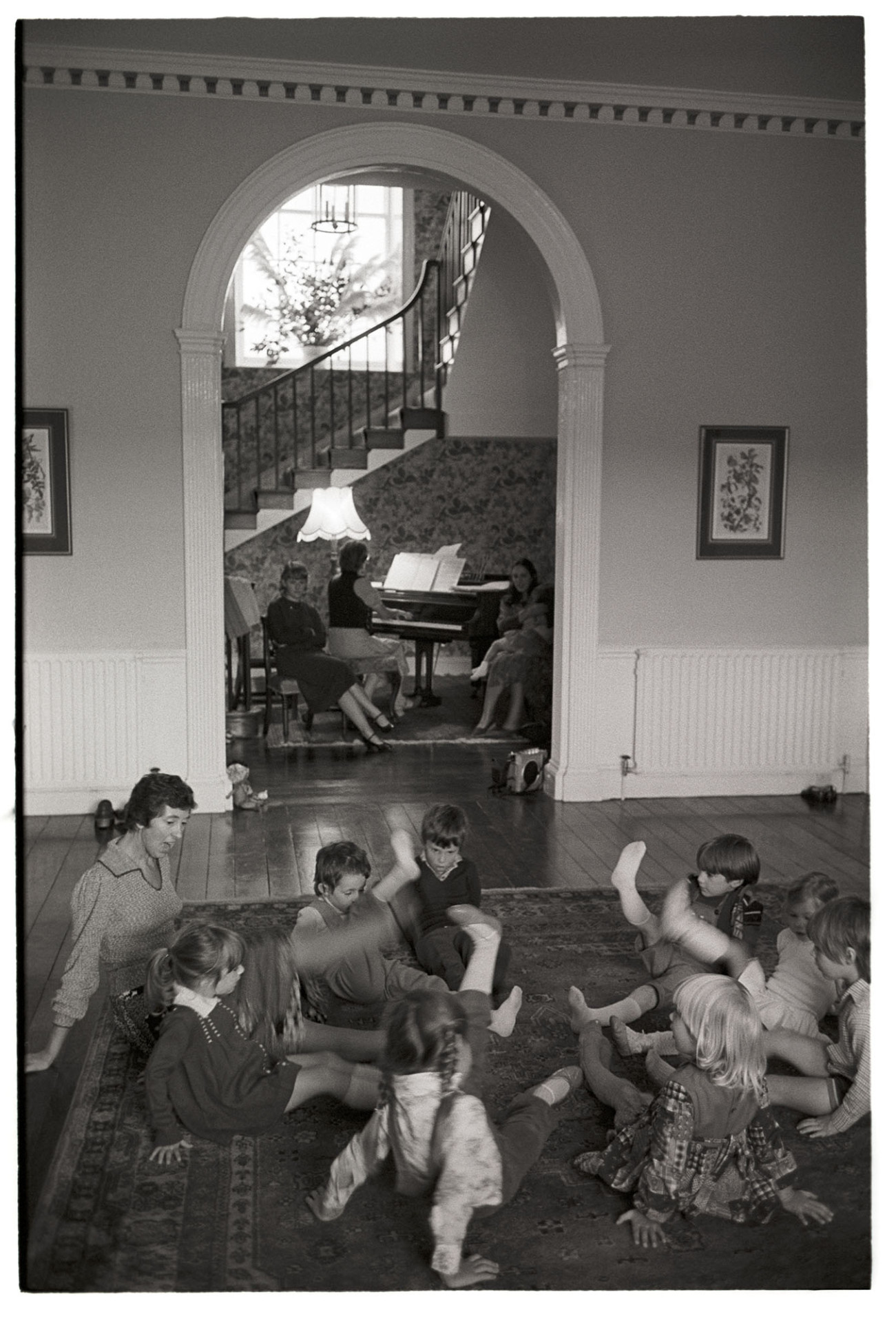 Children's dance class in large country house, teacher. 
[A woman teaching a children's dance class at Marwood House. Through an arched doorway people can be seen watching the dance class by a woman playing a piano.]