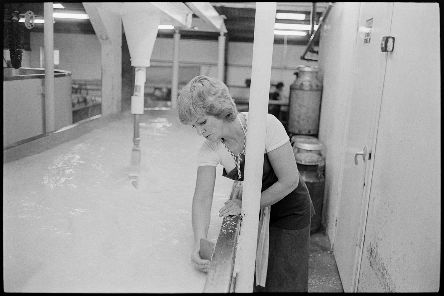 Men and women working in cheese factory, mixing vats, tea break, weighing machine.
[A woman working with liquid in a vat in the cheese factory at Hawkridge Farm, Chawleigh.]