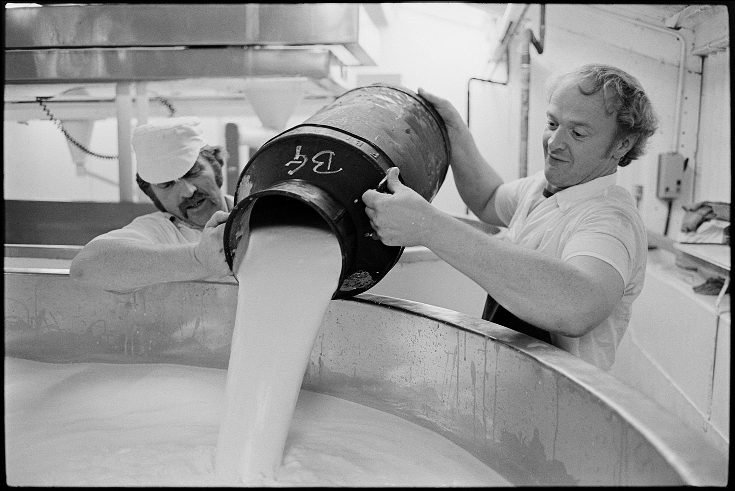 Men and women working in cheese factory, mixing vats, emptying churn into vat. 
[Two men working in the cheese factory at Hawkridge Farm, Chawleigh. They are emptying a churn of milk into a vat.]