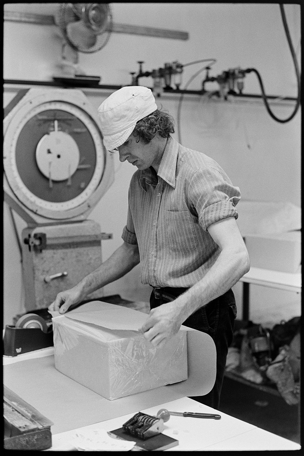 Men and women working in cheese factory, mixing vats, weighing machine. 
[A man wrapping a block of cheese in the cheese factory at Hawkridge Farm, Chawleigh. He is wearing a hat.]
