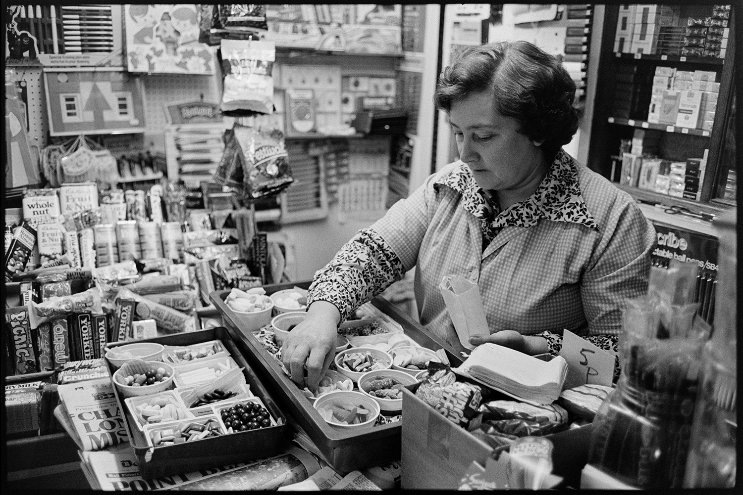 Two women behind counter of shop, sweets and cigarettes on shelf, waiting for children. 
[Joyce Jones sorting pots of sweets in a tray in her shop in Winkleigh Square. Chocolate bars and bags of sweets are displayed on the shelf behind her.]