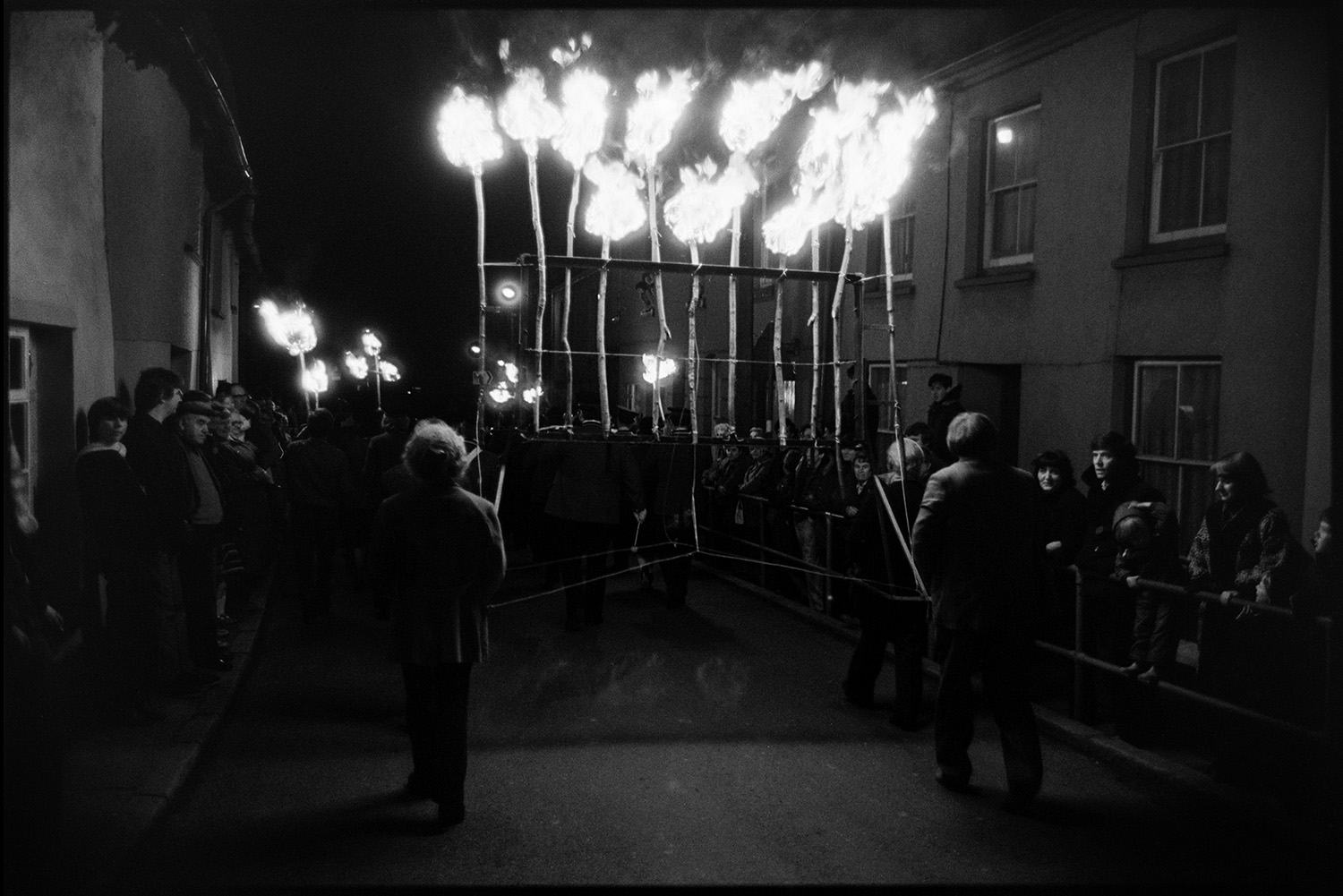Flares and floats at night. <br /> [Men carrying a wooden frame with flaming torches through a street at Hatherleigh Carnival at night. People are lining the street, watching the parade.]
