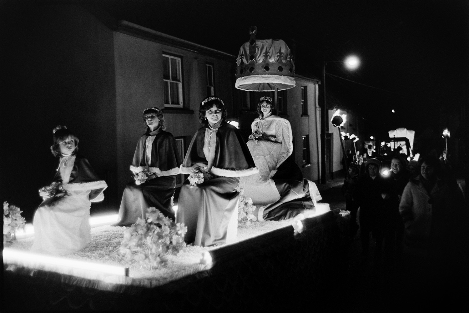 Flares and floats at night. <br /> [The Hatherleigh Carnival Queen and her attendants on a carnival float parading through a street at Hatherleigh Carnival. People carrying flaming torches are following them in the parade.]