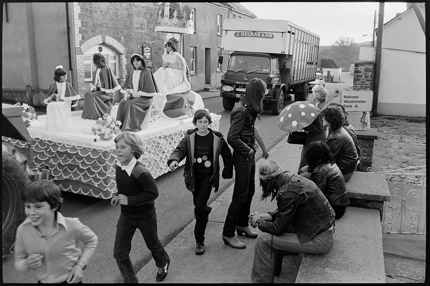 Crowning of Carnival Queen on float, procession through town with onlookers. 
[Teenagers wearing leather jackets sat on a wall watching the Hatherleigh Carnival Queen and her attendants parade past on a carnival float. Three boys are also running past them. A livestock lorry is following the carnival float along the street.]