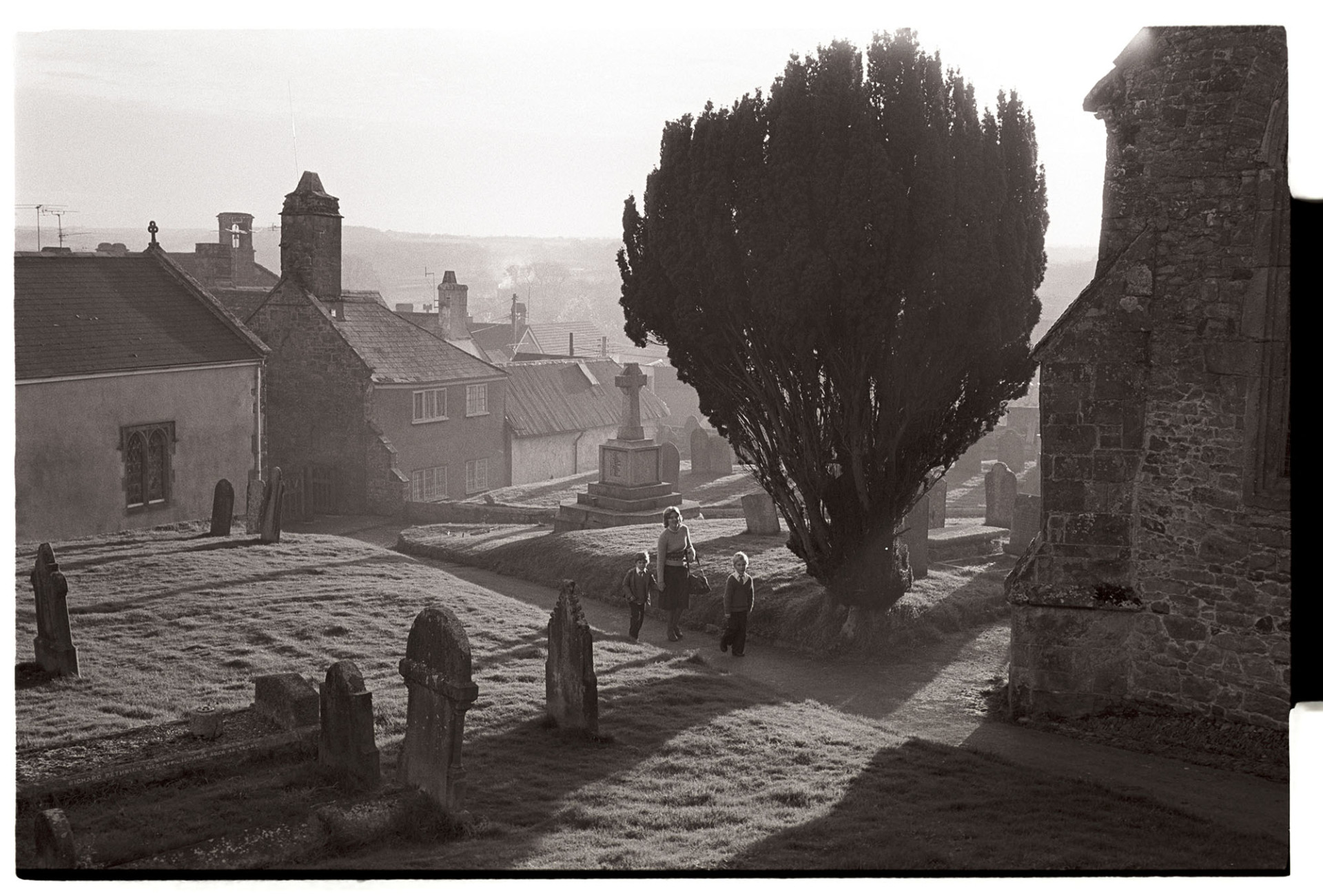 Graveyard, early morning with children going to school. 
[A woman with two children walking through Hatherleigh churchyard on their way to school. Gravestones, a war memorial and a yew tree are all visible in the churchyard.]