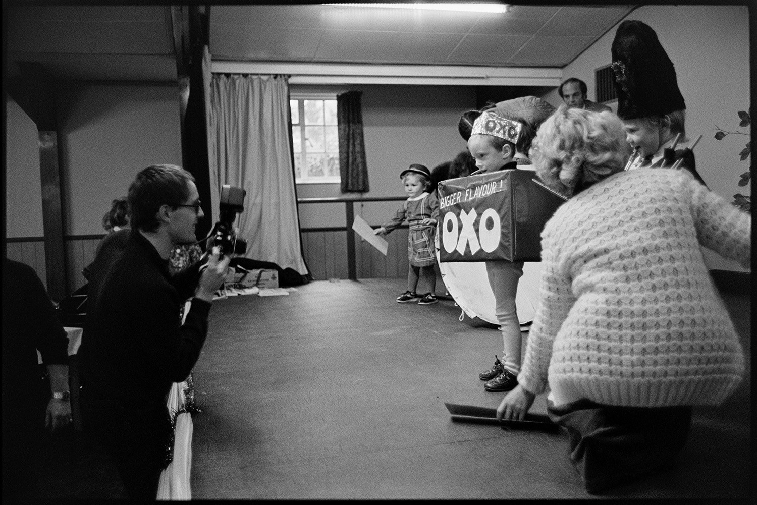 Fancy dress competition in village hall, spectators, queen and crown on cushion. 
[A man photographing young children taking part in a fancy dress competition for Dolton Carnival in Dolton Village Hall. The children are stood on the stage and one child is dressed as an Oxo cube.]
