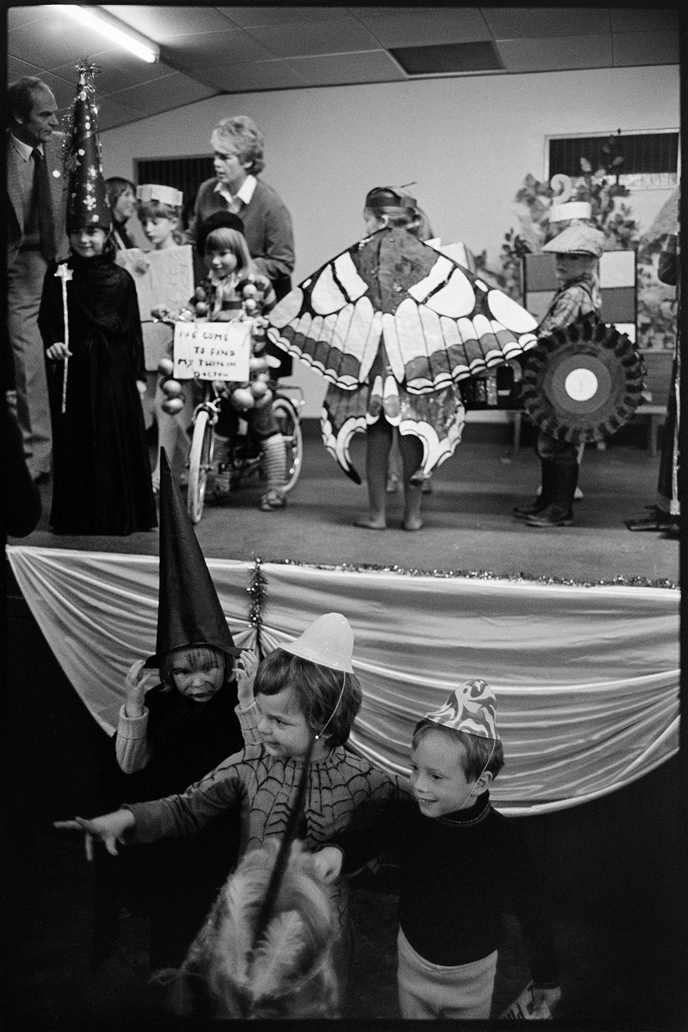 Fancy dress competition in village hall, spectators, queen and crown on cushion. 
[Children taking part in a fancy dress competition for Dolton Carnival in Dolton Village Hall. Costumes include a butterfly, a witch or wizard and a child on a bicycle.]