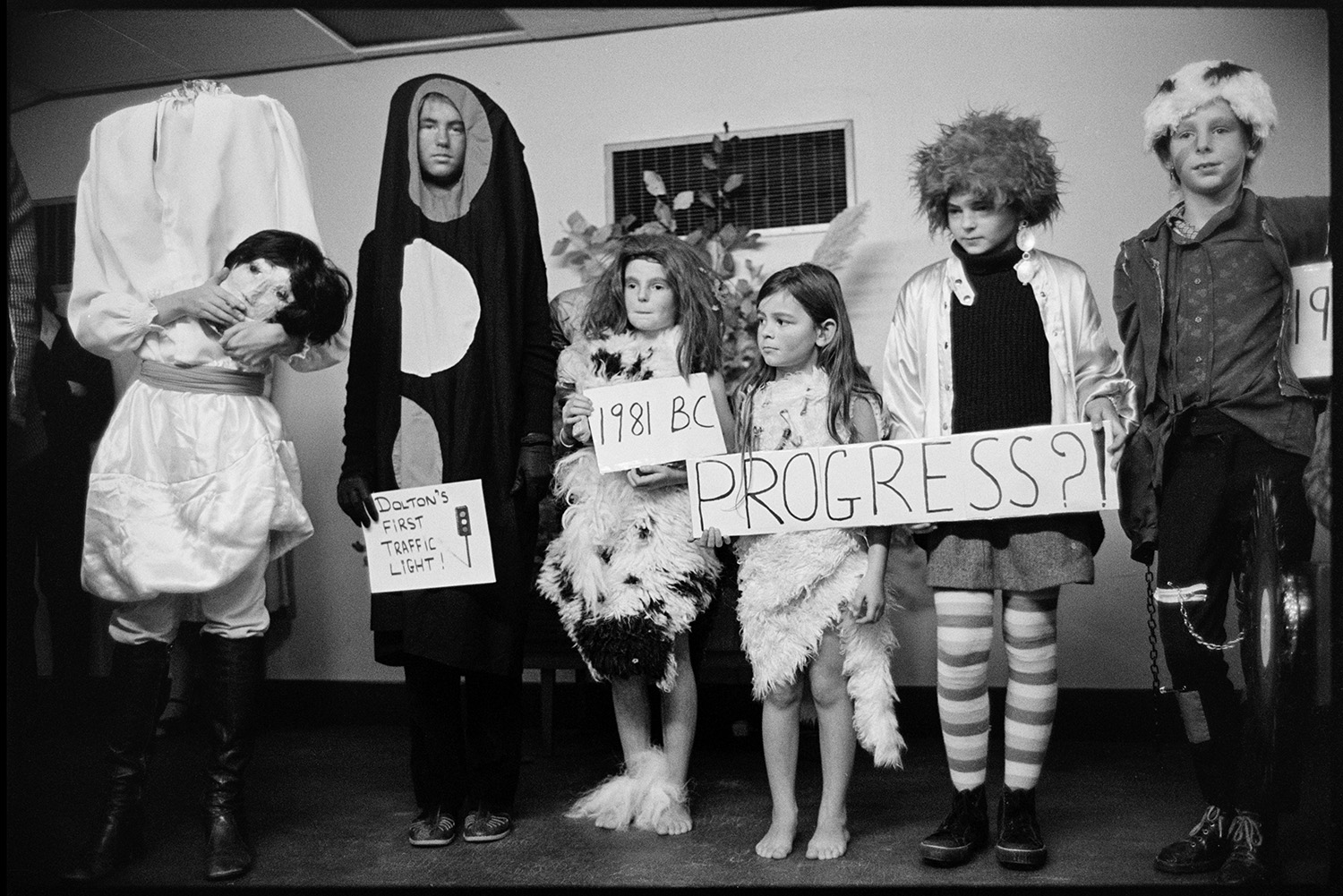 Fancy dress competition in village hall, spectators, queen and crown on cushion. 
[Children at  a fancy dress competition for Dolton Carnvial, in Dolton Village Hall. One child is dressed as a headles sperson and another is a traffic light. Four children are dressed as the stages of man from prehistoric man to the present day, and holding the sing 'Progress?'.]