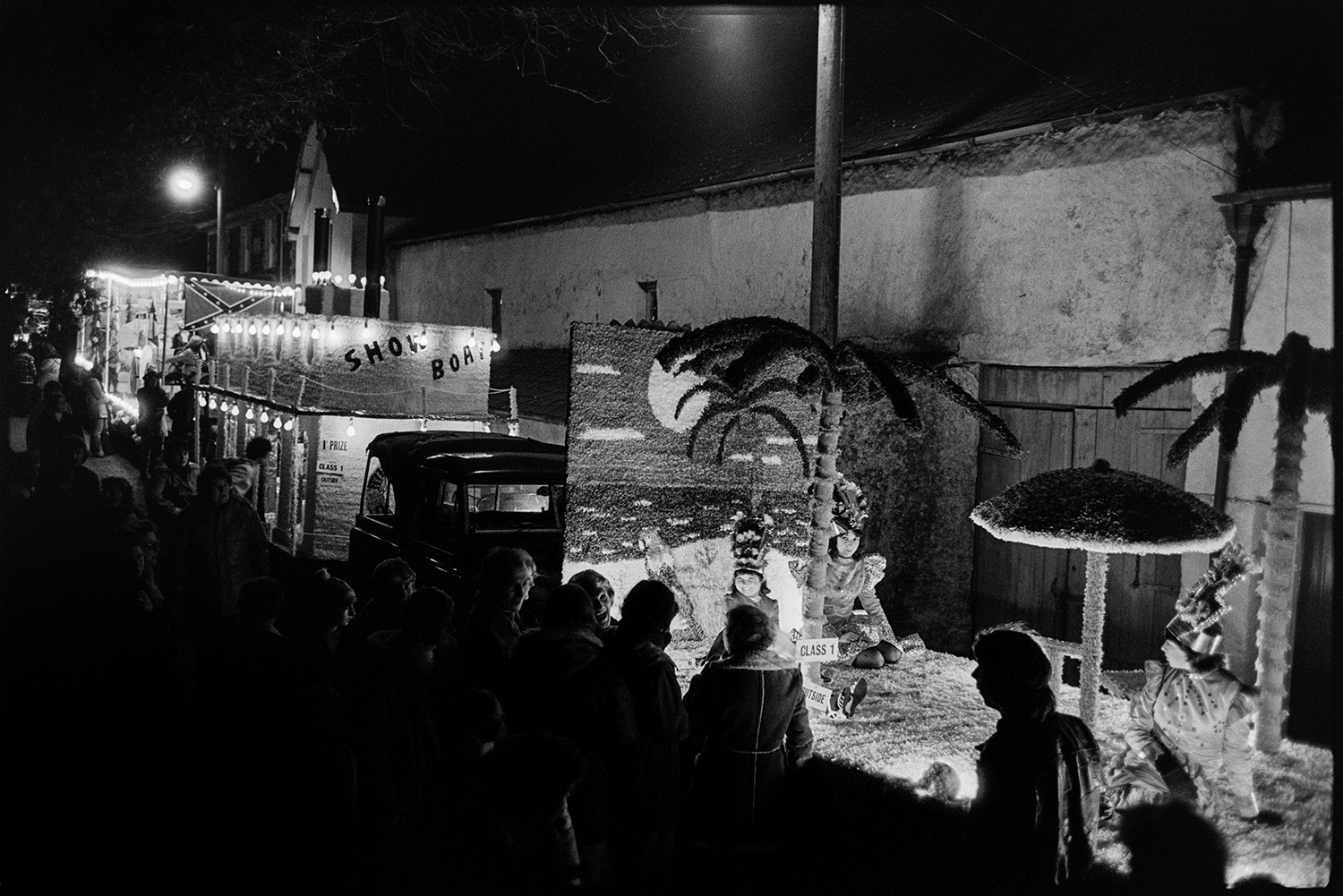 Carnival floats at night people in fancy dress, flares, Bride and Groom. 
[Carnival floats parading through the streets of Dolton at night in Dolton Carnival. People are lining the street watching the parade. The float in the foreground has palm trees and the one behind is titled 'Show Boats'.]