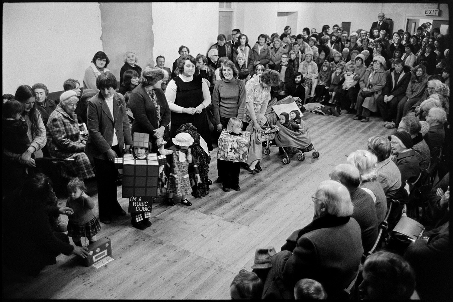 Carnival queen being crowned, fancy dress parade. 
[Children lined up with their mothers at a fancy dress parade in Northlew Village Hall for Northlew Carnival. One child is dressed as a Rubik's cube. An audience is watching the parade.]