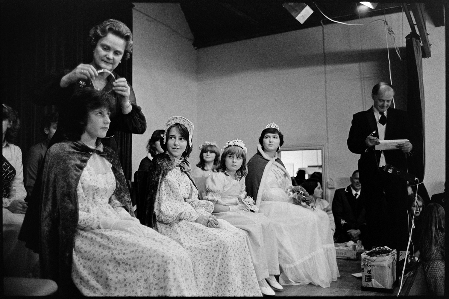 Carnival queen being crowned, fancy dress parade. 
[A woman crowing the Northlew Carnival Queen and Princesses in the village hall.]