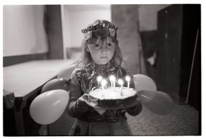 Girl with birthday cake waiting to go on the fancy dress parade by James Ravilious