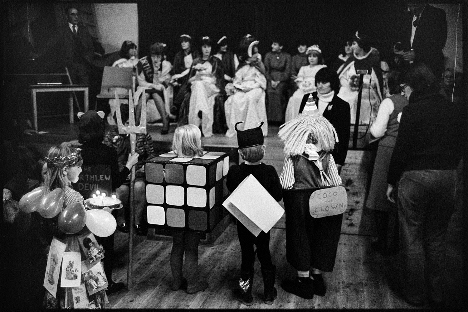 People in fancy dress waiting to go on parade in village hall. 
[Children taking part in a fancy dress parade in Northlew Village Hall for Northlew Carnival. One child is dressed as a Rubik's cube and another as Coco the Clown. Another girl is carrying a cake with candles. The carnival Queen and attendants are watching in the background.]