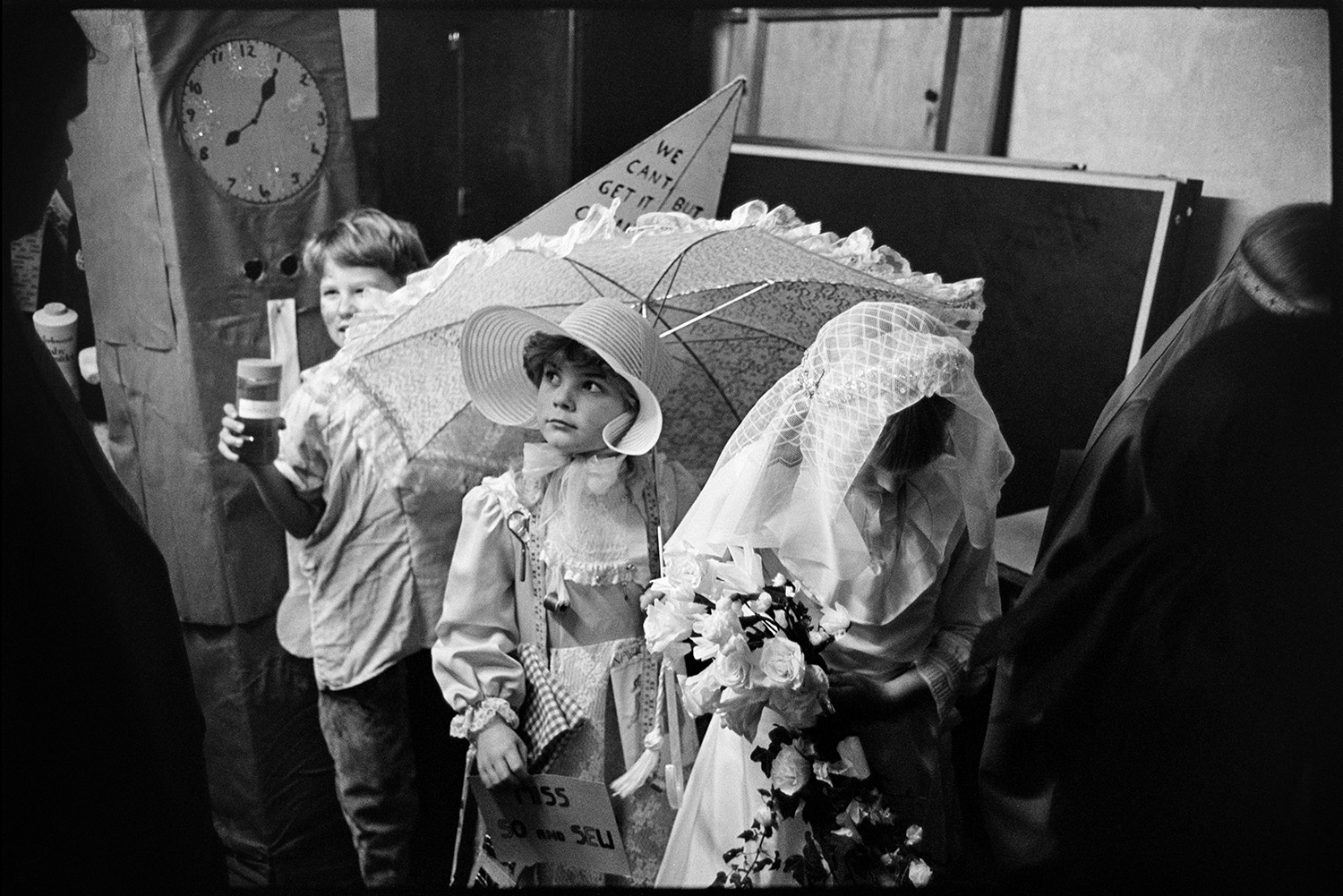 People in fancy dress waiting to go on parade in village hall. 
[Children waiting to go on a fancy dress parade in Northlew Village Hall for Northlew Carnival. One child is dressed as a bride, another girl is carrying a parasol and a child at the back is wearing a grandfather clock costume.]