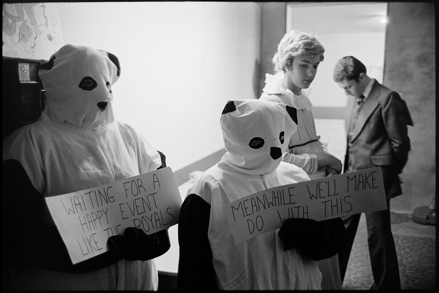 People in fancy dress waiting to go on parade in village hall. 
[Two people dressed as pandas for a fancy dress parade in Northlew Village Hall for Northlew Carnival. They are each holding a placard which reads 'Waiting For A Happy Event Like The Royals' and 'Meanwhile We'll Make Do With This'.]