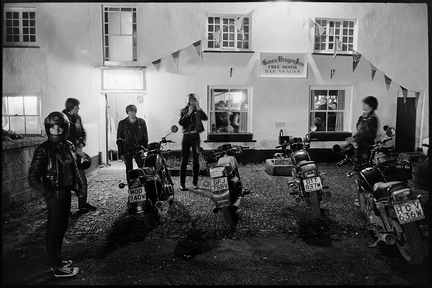 Motorbikes parked outside pub at night, carnival night. 
[Young men stood by their motorbikes outside the Green Dragon Inn in Northlew on the night of the village carnival. The pub is decorated with bunting.]