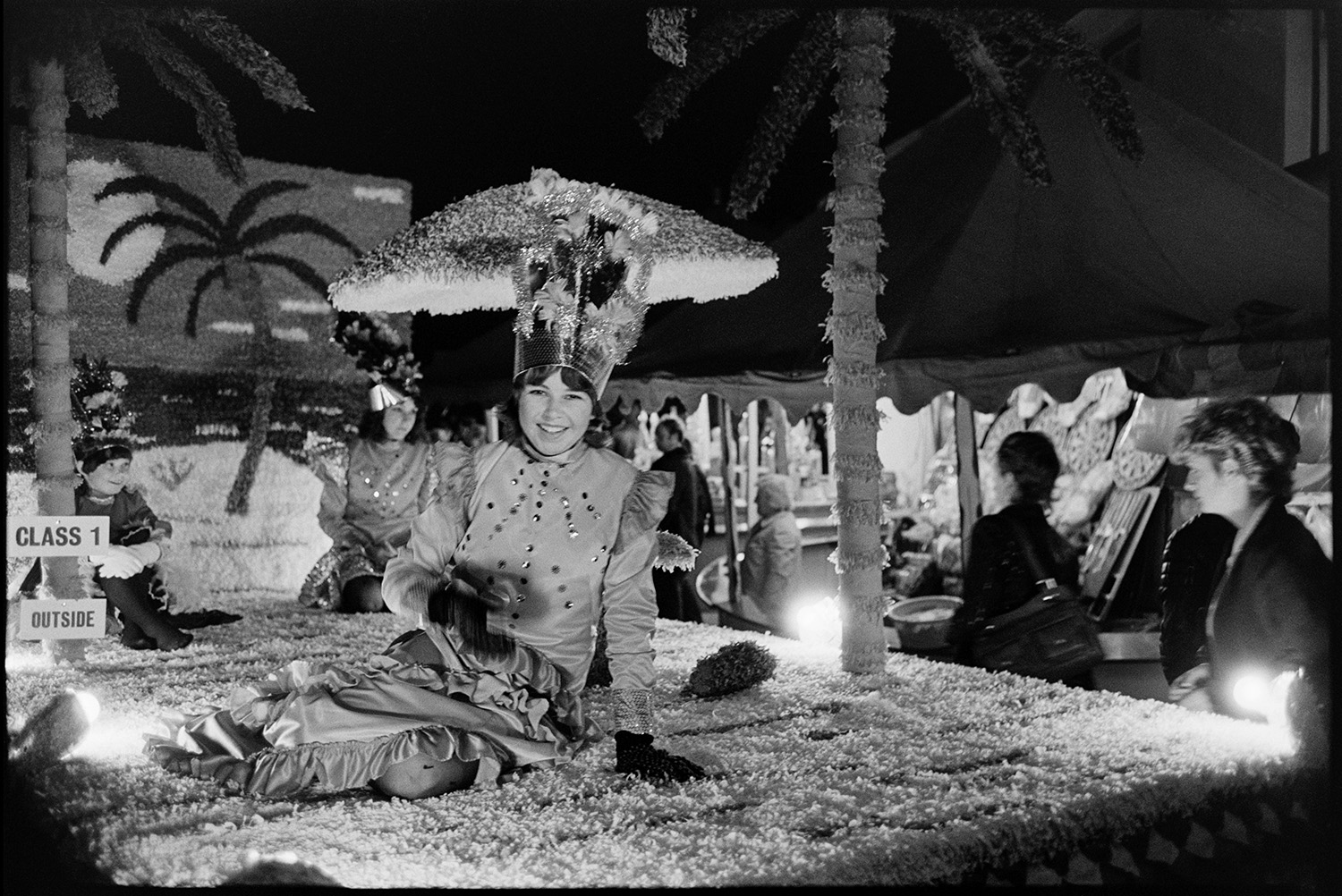 Girls, fancy dress on float at night. 
[Girls in fancy dress on a carnival float with palm trees in Dolton Carnival at night.]