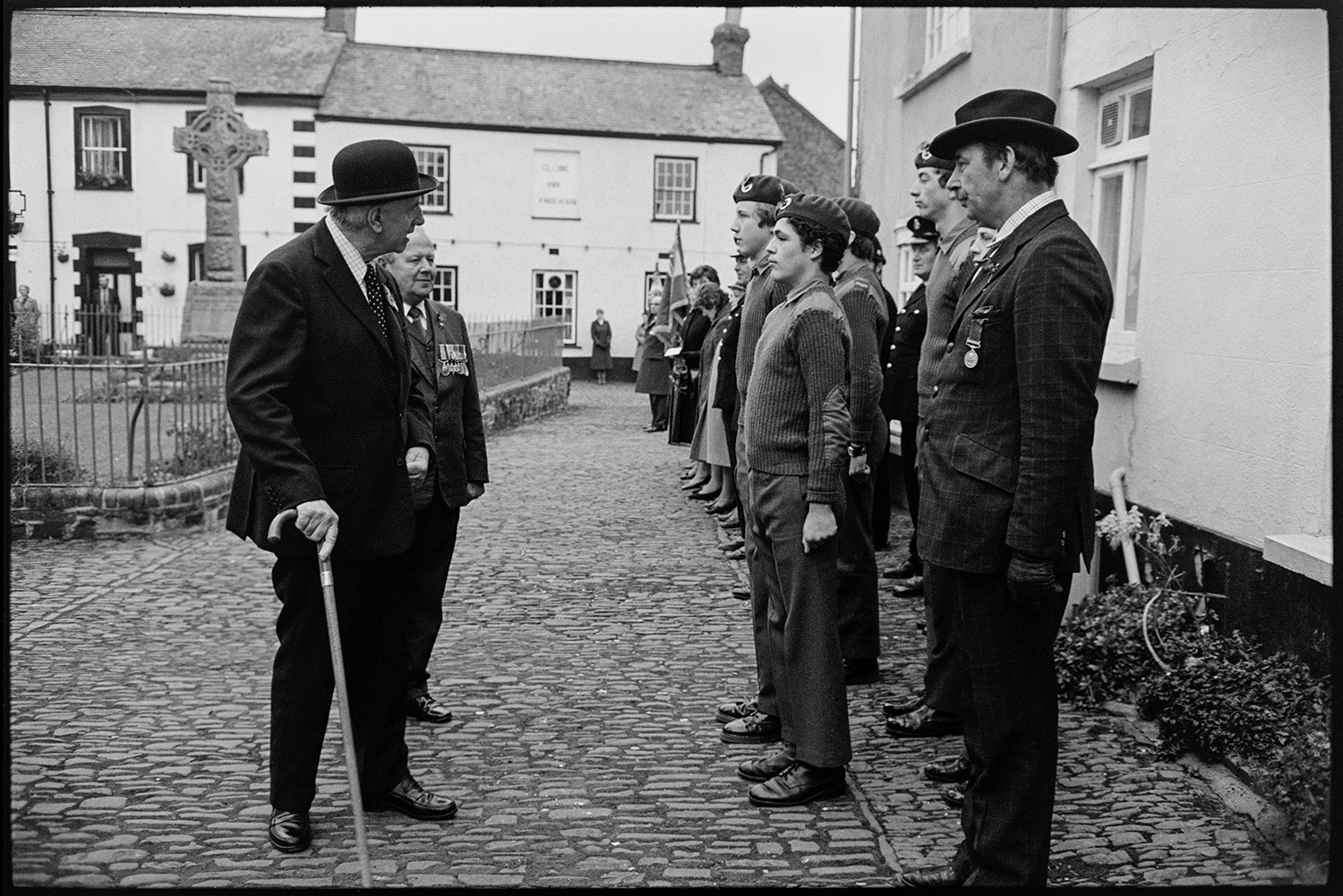 Armistice Day, inspection and parade to church, both women and men. Scouts and brownies. 
[Two men, one wearing medals and another wearing a bowler hat and using a walking stick, inspecting cadets lined up on a cobbled street by Chulmleigh war memorial on Remembrance Day.]