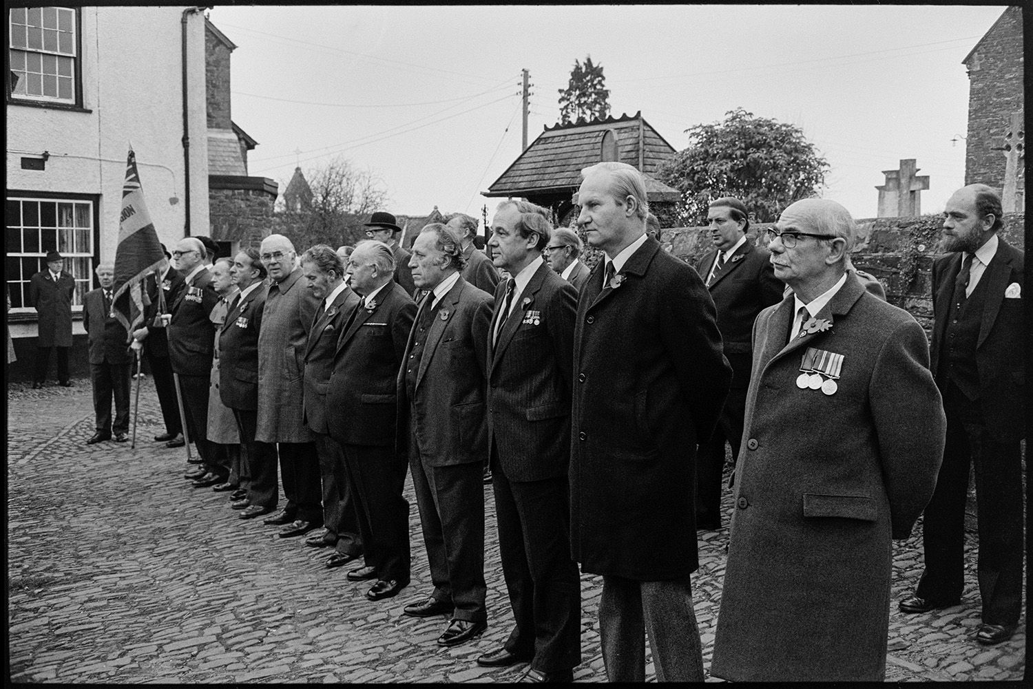 Armistice Day, inspection and parade to church, both women and men. Scouts and brownies. 
[Men, possibly ex-servicemen, lined up on a cobbled street by the Chulmleigh war memorial for Remembrance Day. Some of them are wearing medals.]