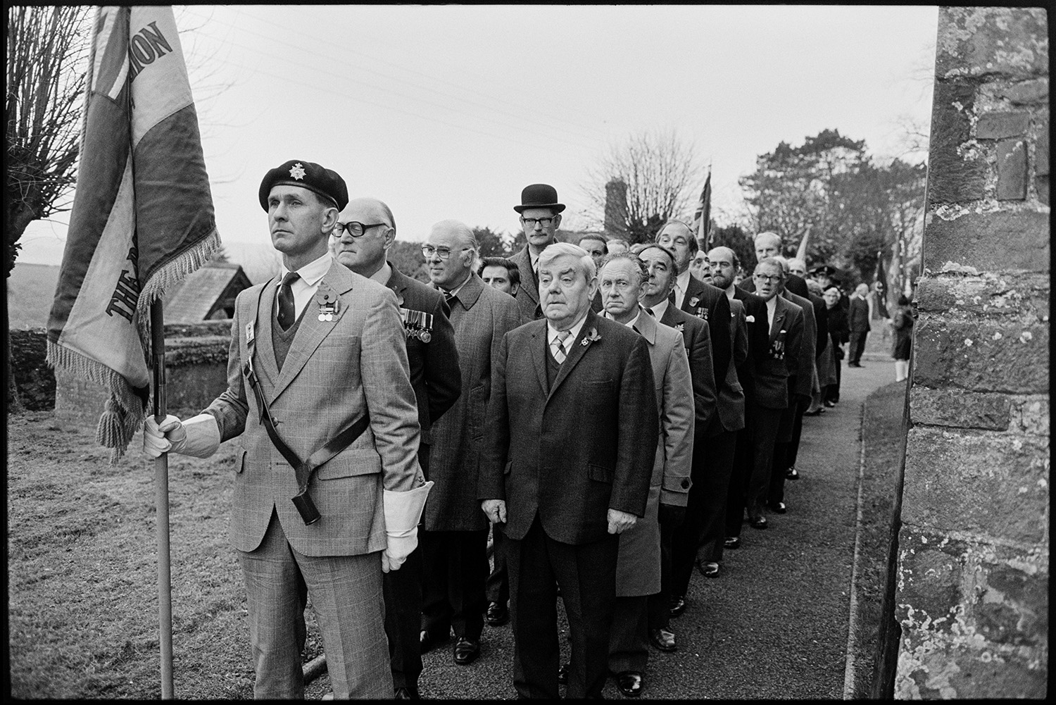 Armistice Day, church, parade line up, chat afterwards with officers in bowler hats, medals. 
[Men, possibly ex-servicemen, lined up outside Chulmleigh Church for a Remembrance Day parade. One man is wearing a bowler hat. Some of them are wearing medals. The man leading the parade is carrying a British Legion flag.]