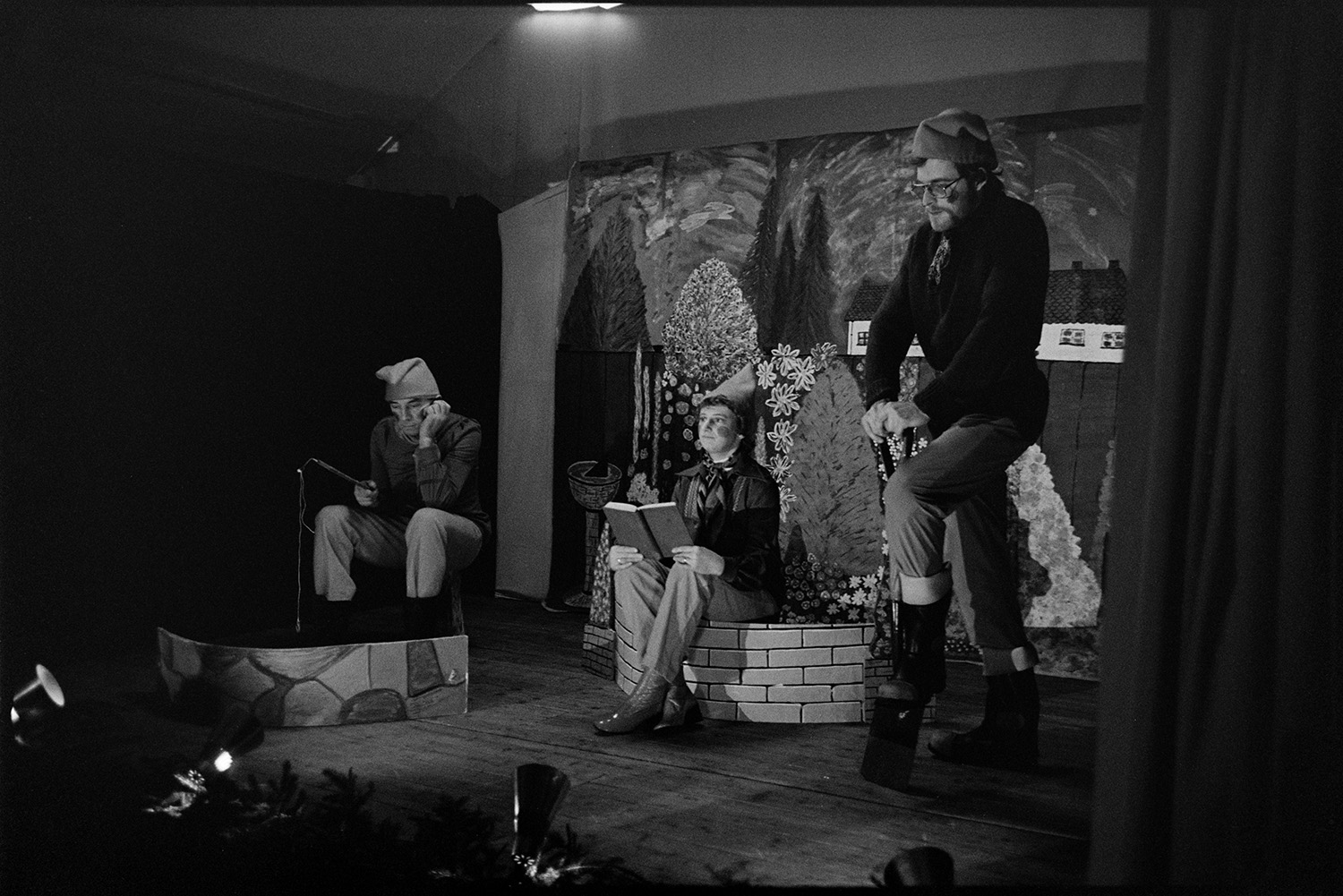 Christmas entertainment in village hall, singing and dance, short plays.
[Three people dressed as gnomes on stage at High Bickington village hall. They are performing a short play for Christmas entertainment. One is fishing, one is reading a book and the other is digging with a spade.]